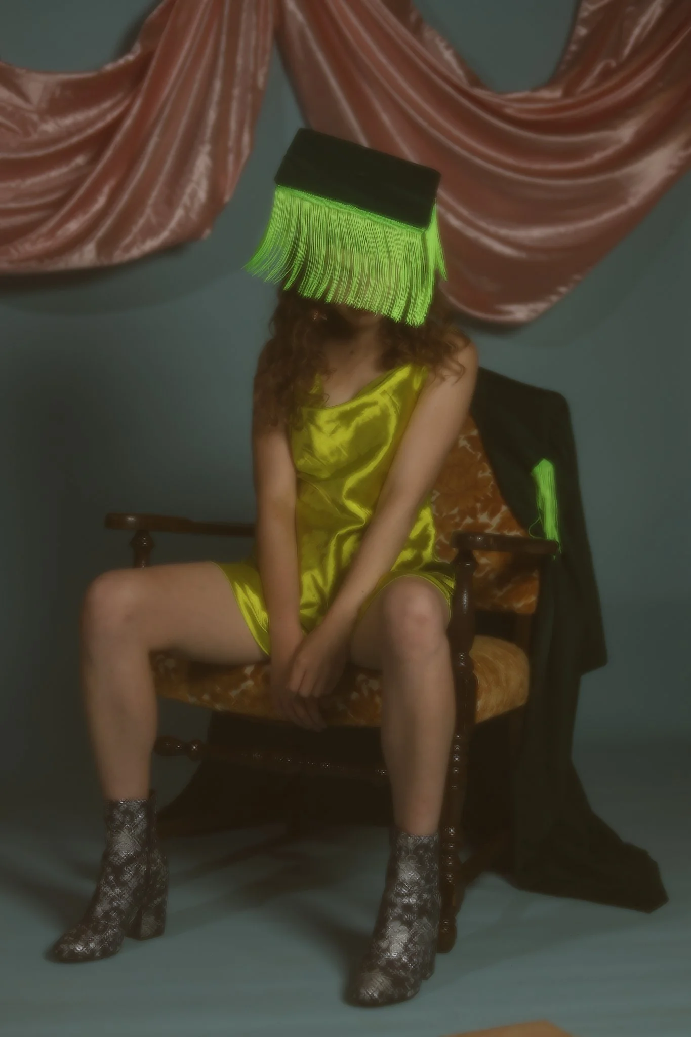 A person with curly hair wearing a yellow satin dress is sitting on a vintage chair with a person wearing snakeskin boots standing in front of them. The person's face is covered by a green glow from a broomstick placed vertically in front of their face. The background features pink velvet curtains.