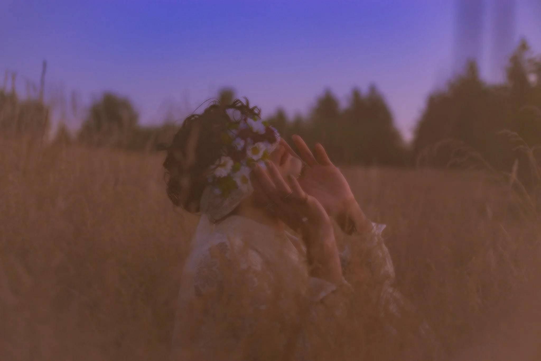 A person with curly hair wearing a flower eye mask and light-colored clothing, standing in a field during sunset or sunrise, with a clear sky in the background.