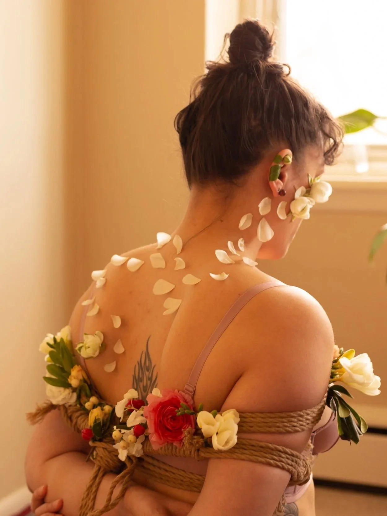 A woman with short hair tied in a bun, decorated with flowers, is seen from the back and side, wrapped with a rope and adorned with flower petals, a tattoo, and flowers on her arms and shoulder. She is near a window with bright natural light.