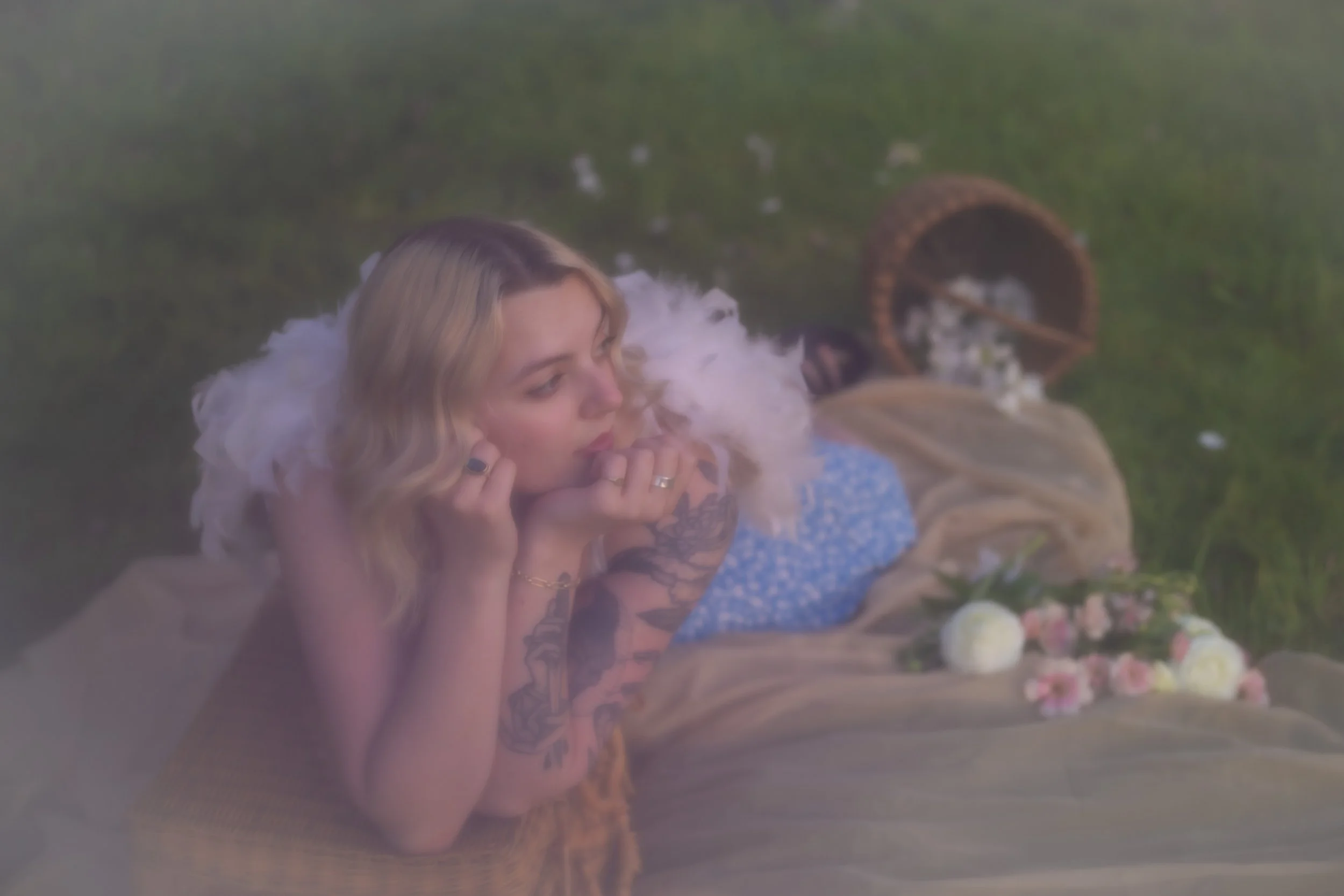 A woman with blonde hair and tattoos on her arm lies on the grass with her eyes closed, her hand resting on her face. She is near a basket spilled with white flower petals and colored flowers.