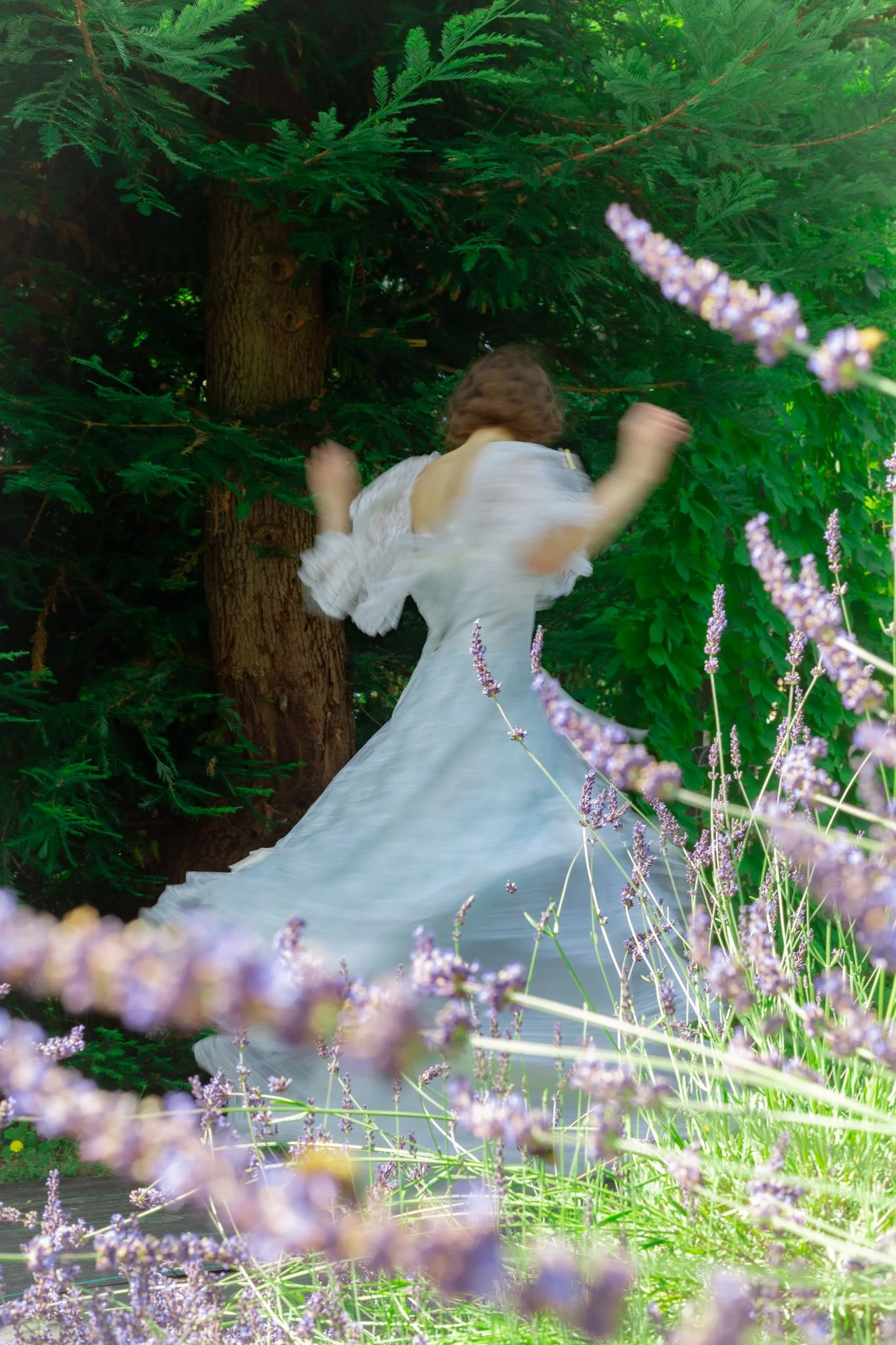 A woman in a white dress with puffed sleeves running through a lush green garden surrounded by purple flowers and tall trees.