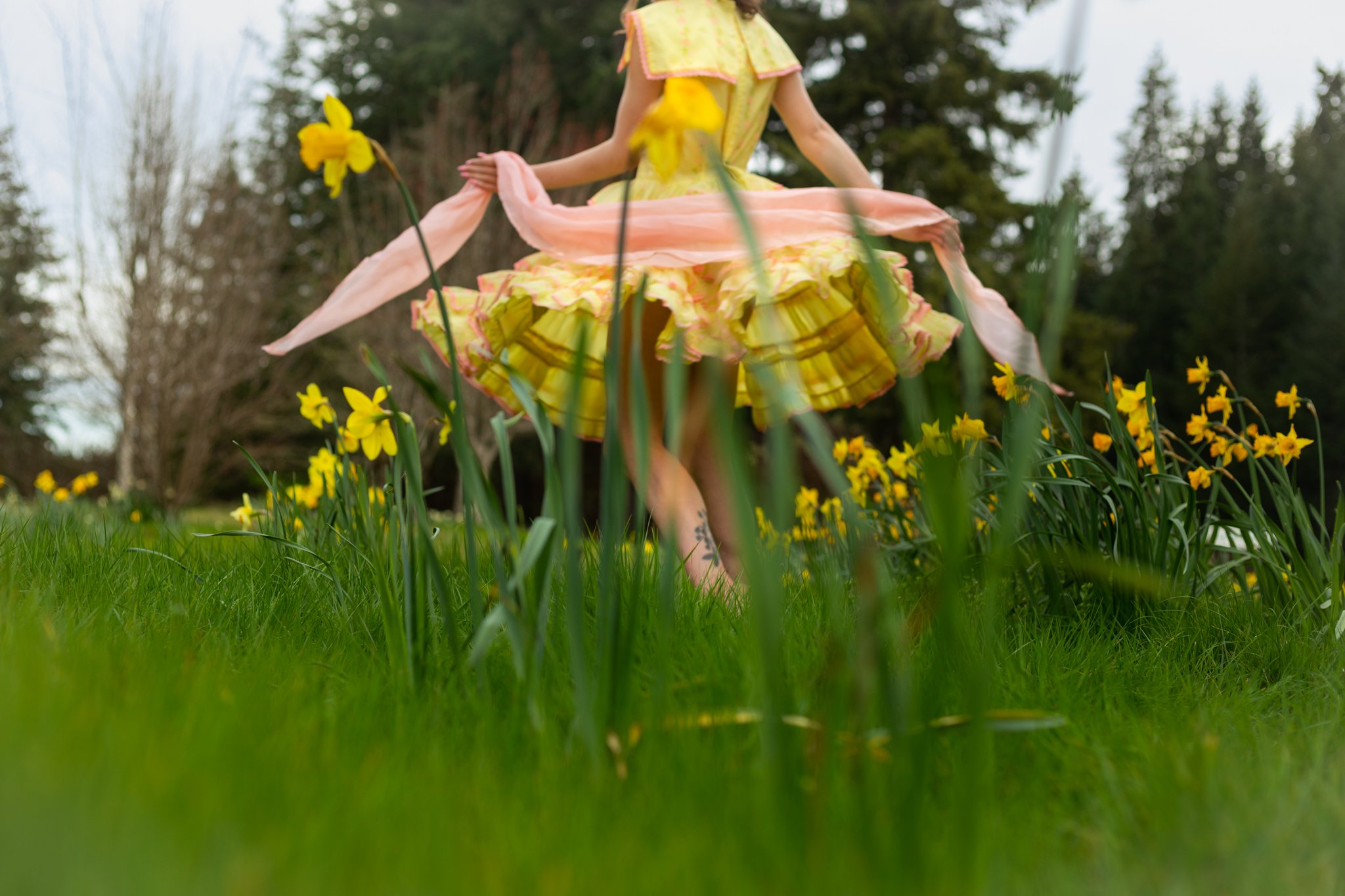 A person wearing a yellow dress with pink and green accents, sitting outdoors among yellow flowers and tall grass, with trees and cloudy sky in the background.