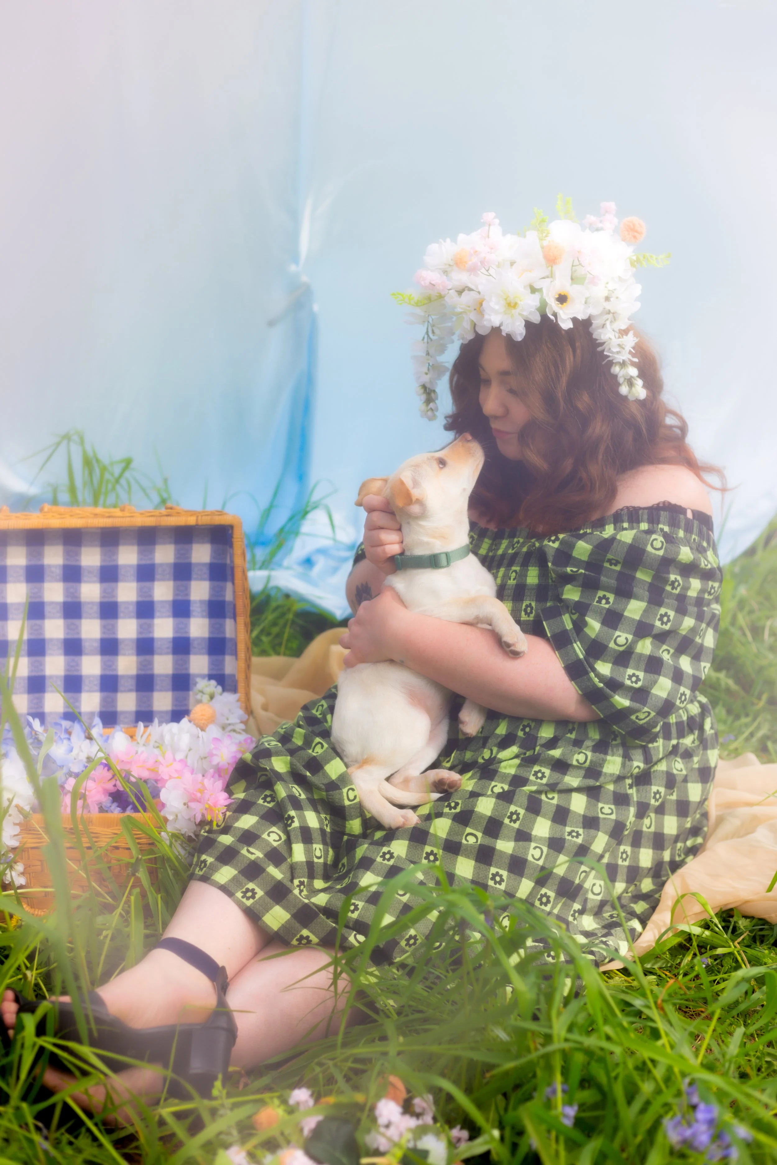 A woman with curly red hair wearing a green checkered dress and a large floral crown holding a small yellow puppy in a grassy outdoor setting. There are flowers and a wicker basket nearby.