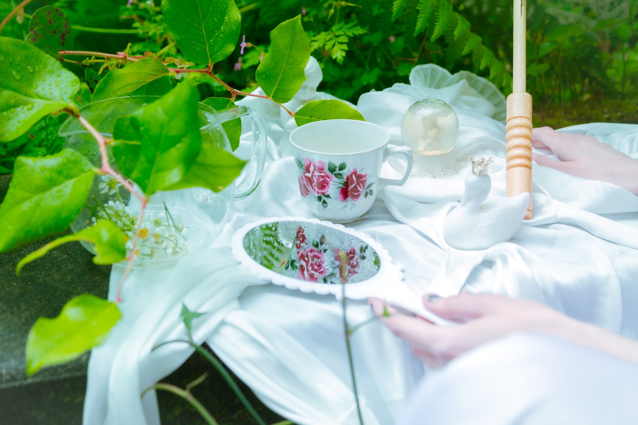 A garden table with a floral teacup, a glass orb, a small silver ring, and gardening tools, surrounded by green leaves.