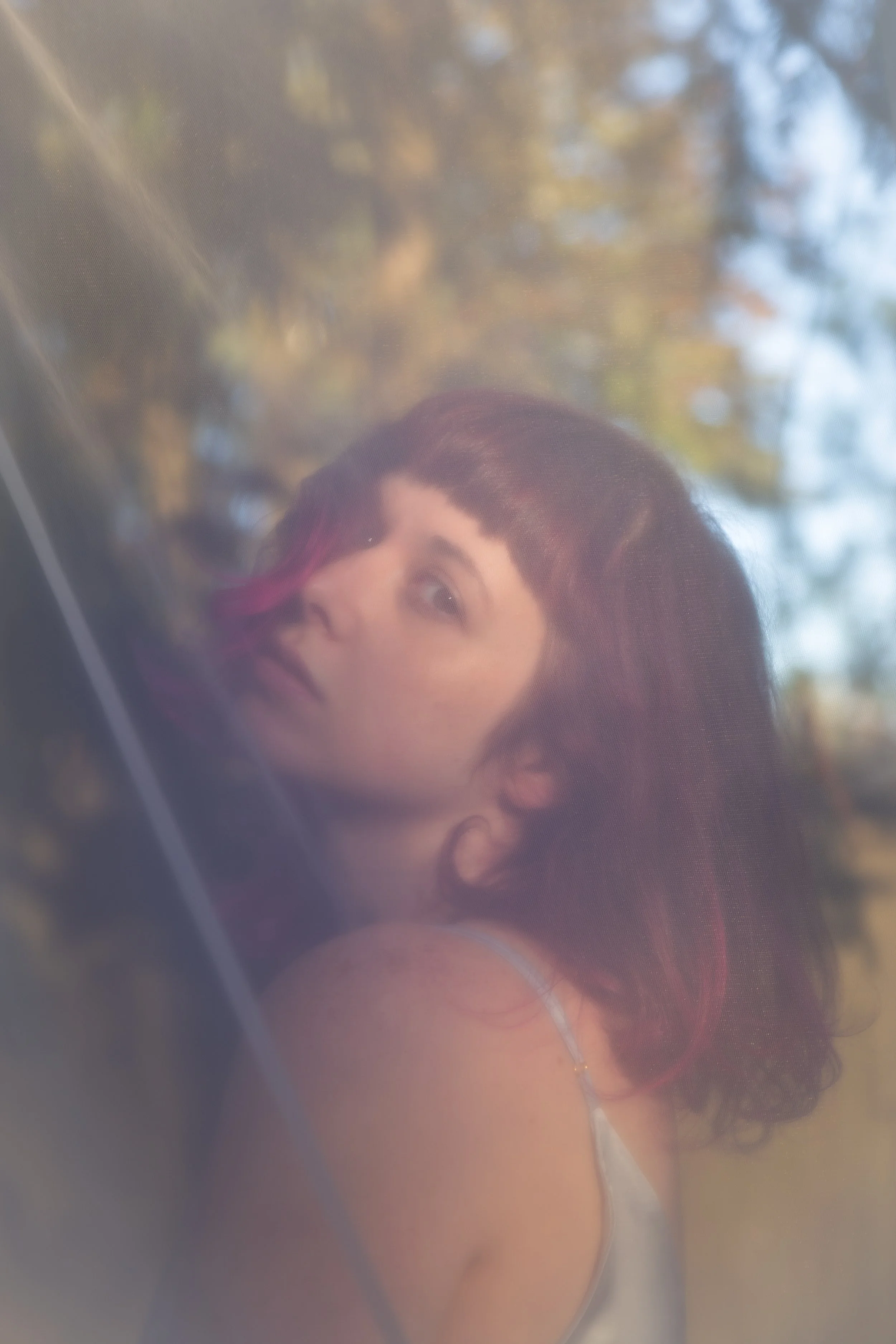 A young woman with curly dark hair and a small pink streak near her forehead. The background shows blurred trees with autumn-colored leaves.