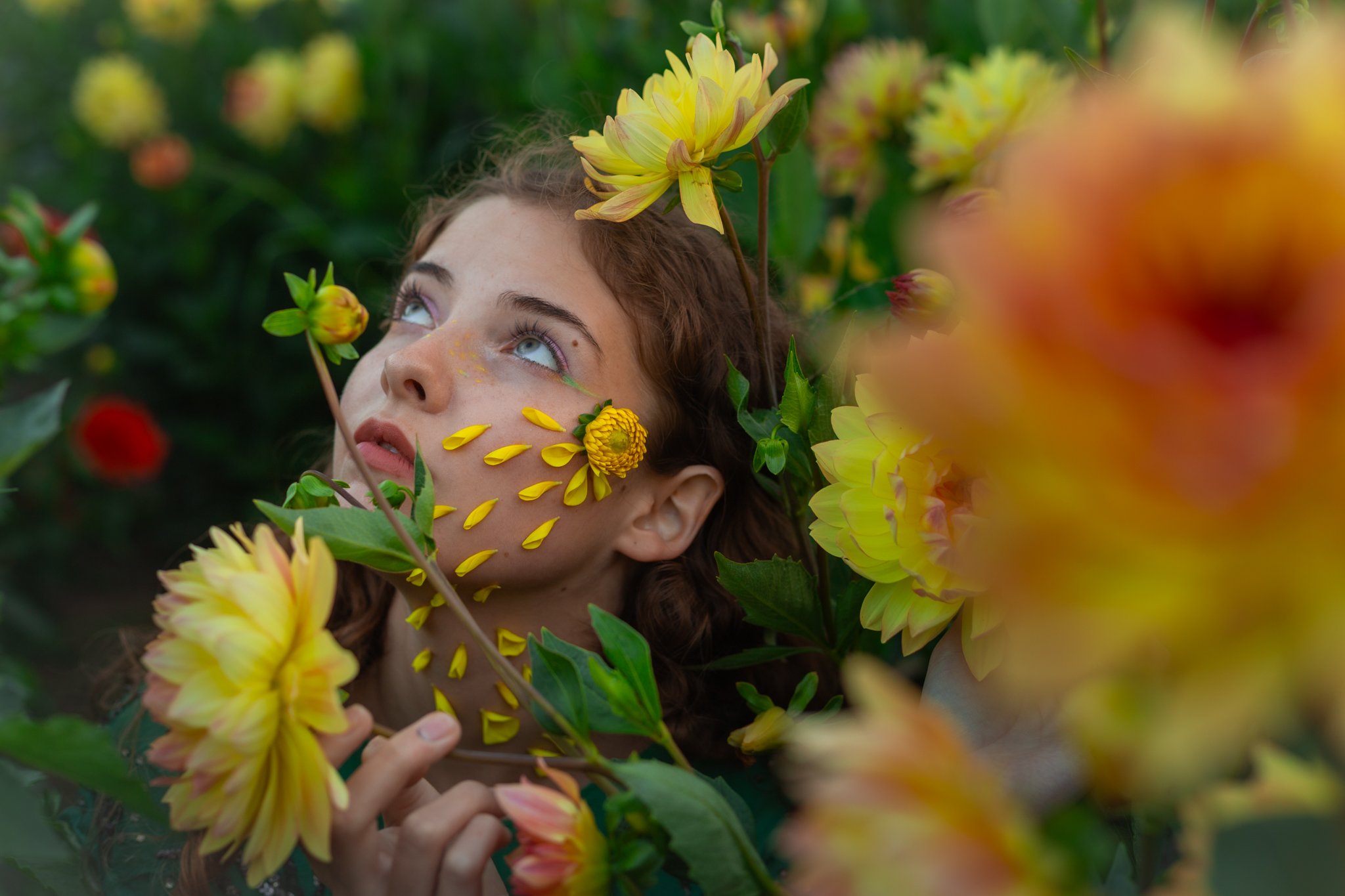 A young woman with curly hair and blue eyes surrounded by yellow flowers; some flowers and petals are on her face and she is looking upwards with her hand gently touching a flower.