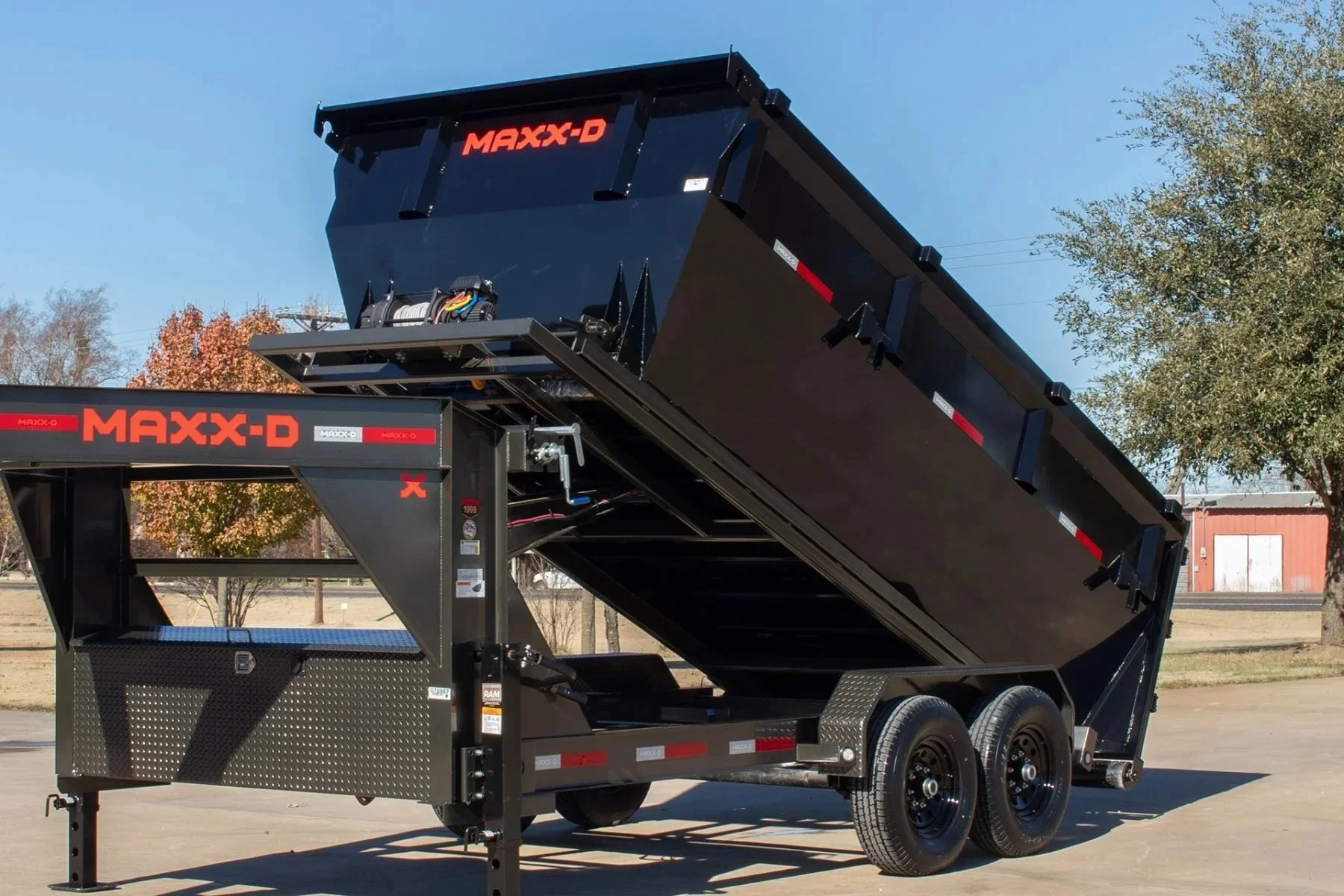 A black MAXX-D dump trailer with the bed raised, showing hydraulic components, parked outdoors on pavement with trees and a red building in the background.