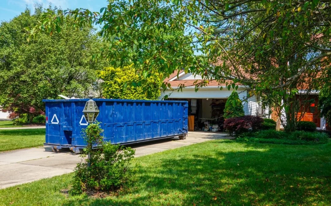 Blue dumpster on driveway in front of a house with a garage, surrounded by green trees and grass.