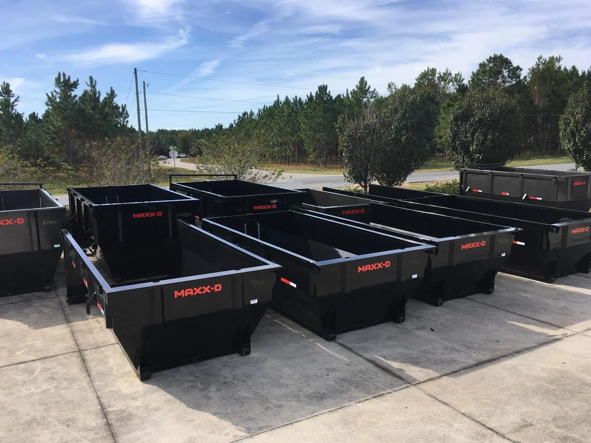 Multiple black MAXX-D trailers lined up on a concrete surface with trees and power lines in the background under a partly cloudy sky.