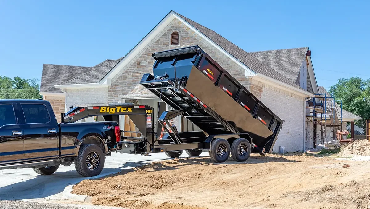 A black pickup truck with a Big Tex dump trailer attached, lifting its bed at a construction site in front of a house under construction with scaffolding and unfinished walls.