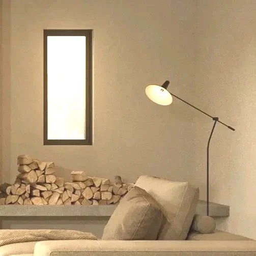 Interior of a minimalist living room with stacked firewood, a beige pillow on a sofa, a tall narrow window, and a modern floor lamp with an adjustable arm.