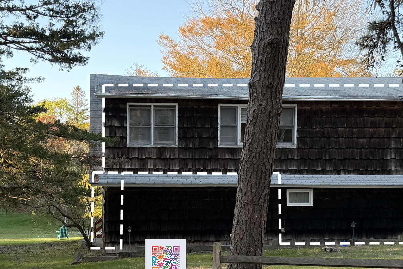 A two-story house with dark wooden shingles, partially obscured by a large tree in the front yard, with a background of trees with autumn-colored leaves.