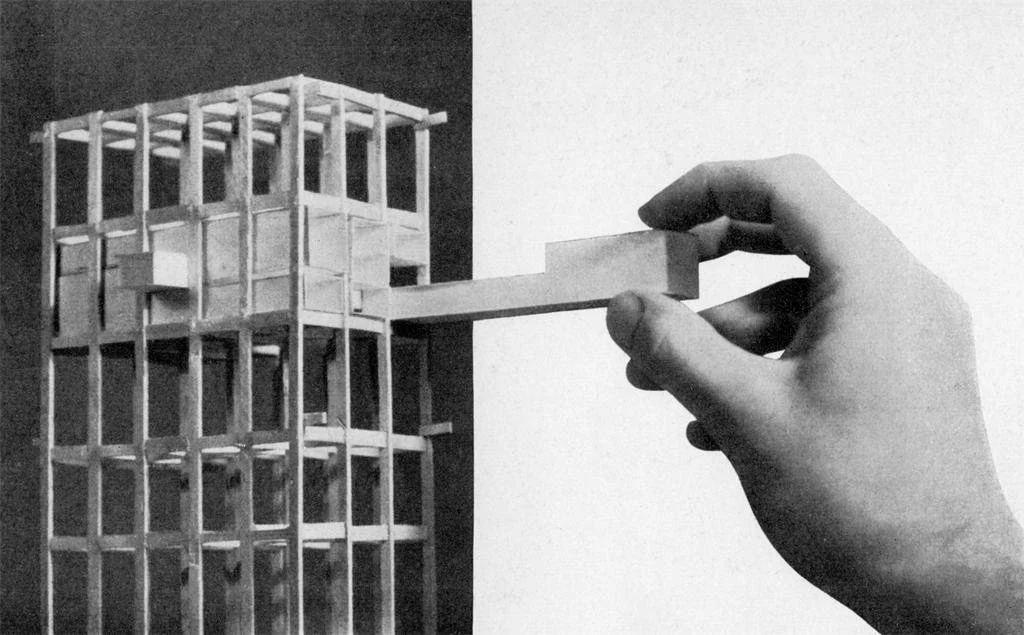 A black and white photo of a hand holding a small wooden block, with a wooden model building in the background.