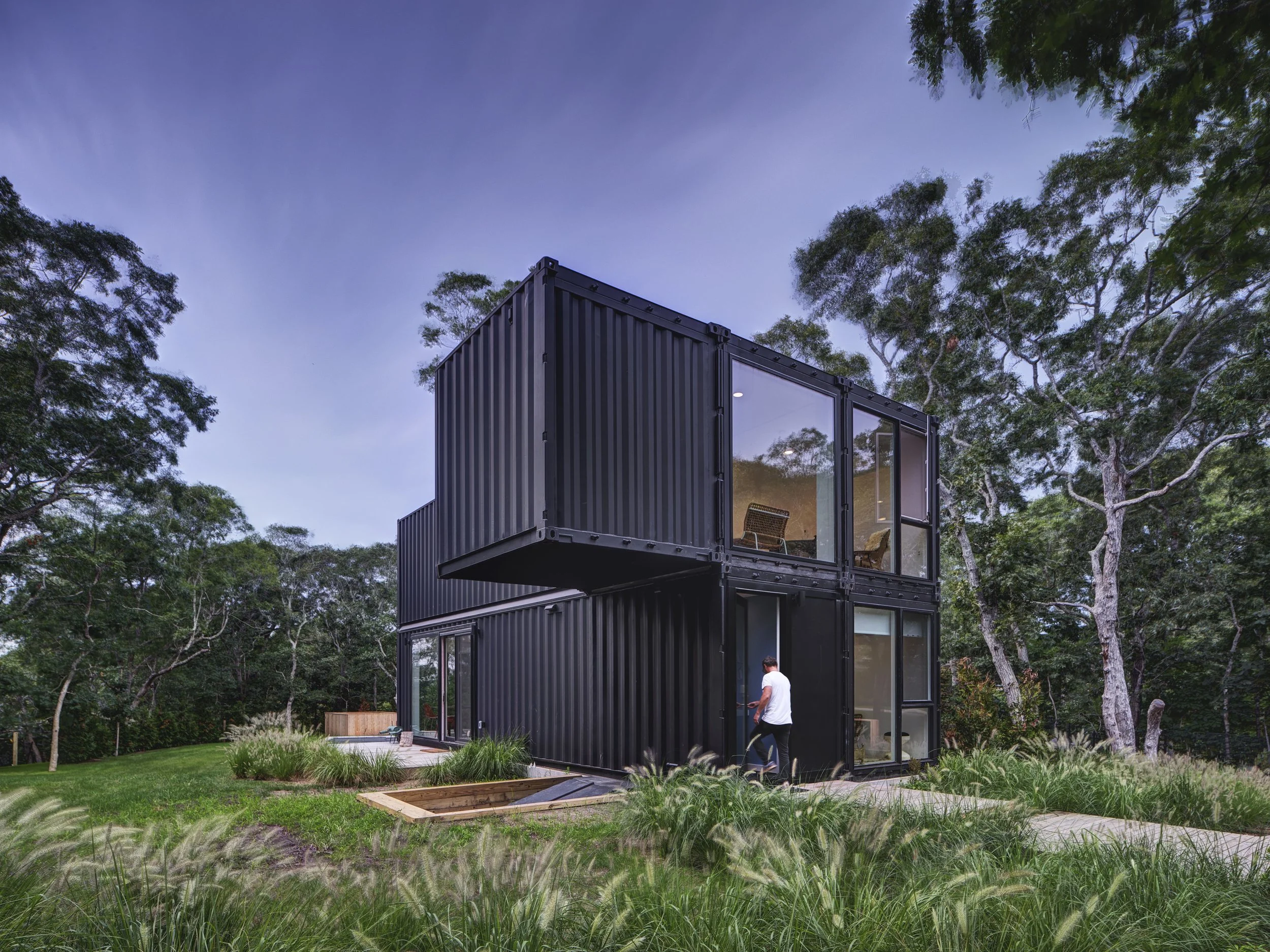 Modern black container home with large glass windows, surrounded by trees and a grassy yard, during dusk.