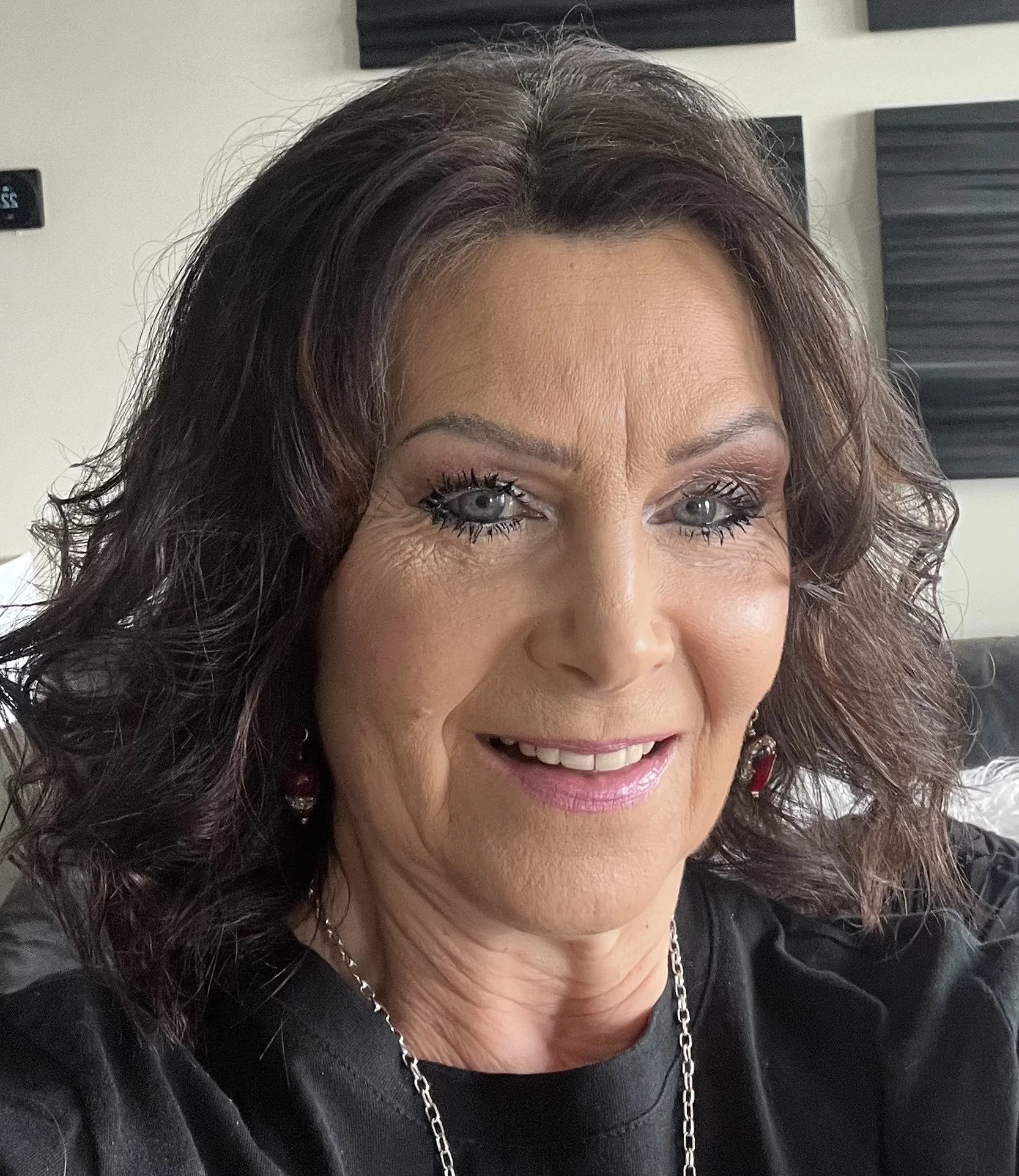 Close-up of a middle-aged woman with curly dark brown hair and blueeyes, smiling, wearing a silver chain necklace.