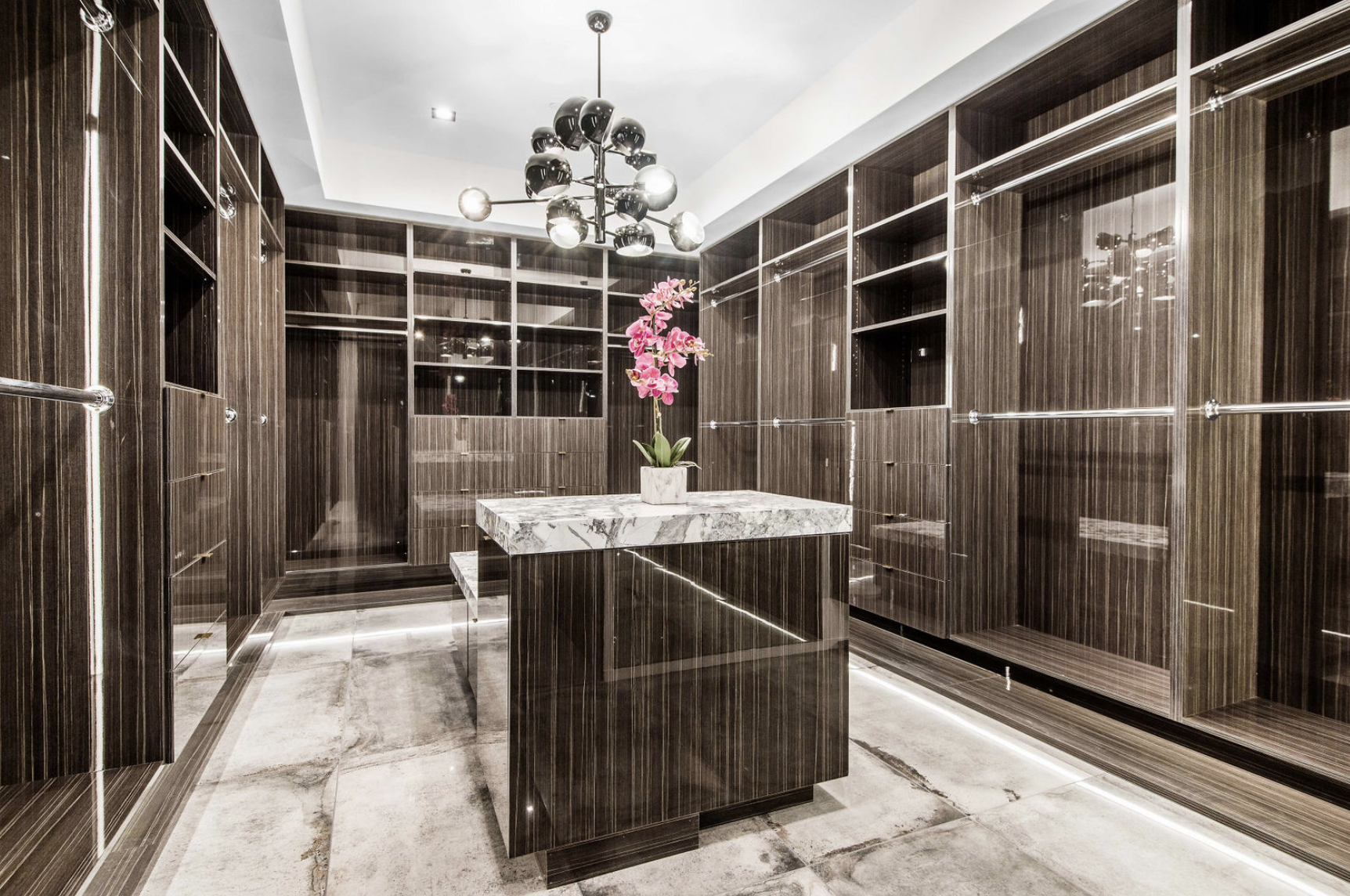 A walk-in closet with dark wood finished shelving and a central marble-top island with a pink orchid plant. The ceiling features a modern black chandelier with multiple globes, and the floor is tiled in light and dark shades.