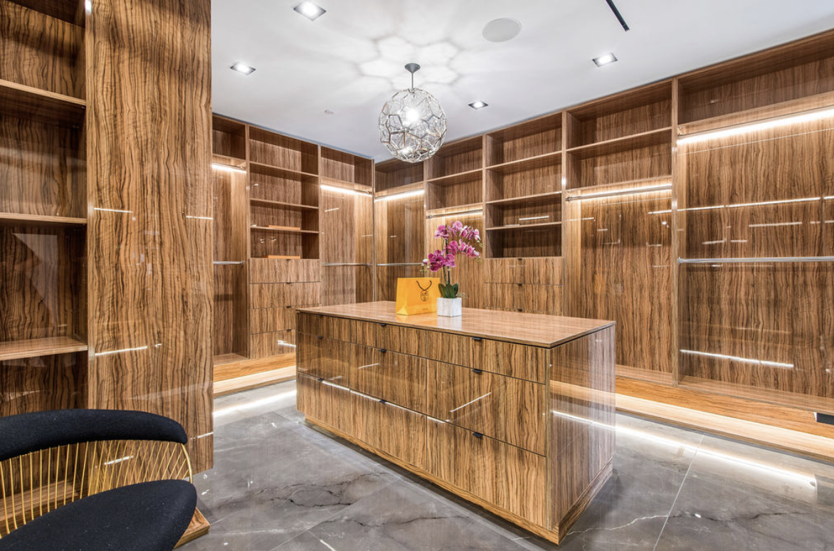 A spacious walk-in closet with wooden shelves and drawers, a central wooden island with a pink orchid and yellow shopping bag on top, modern lighting, and a marble floor.