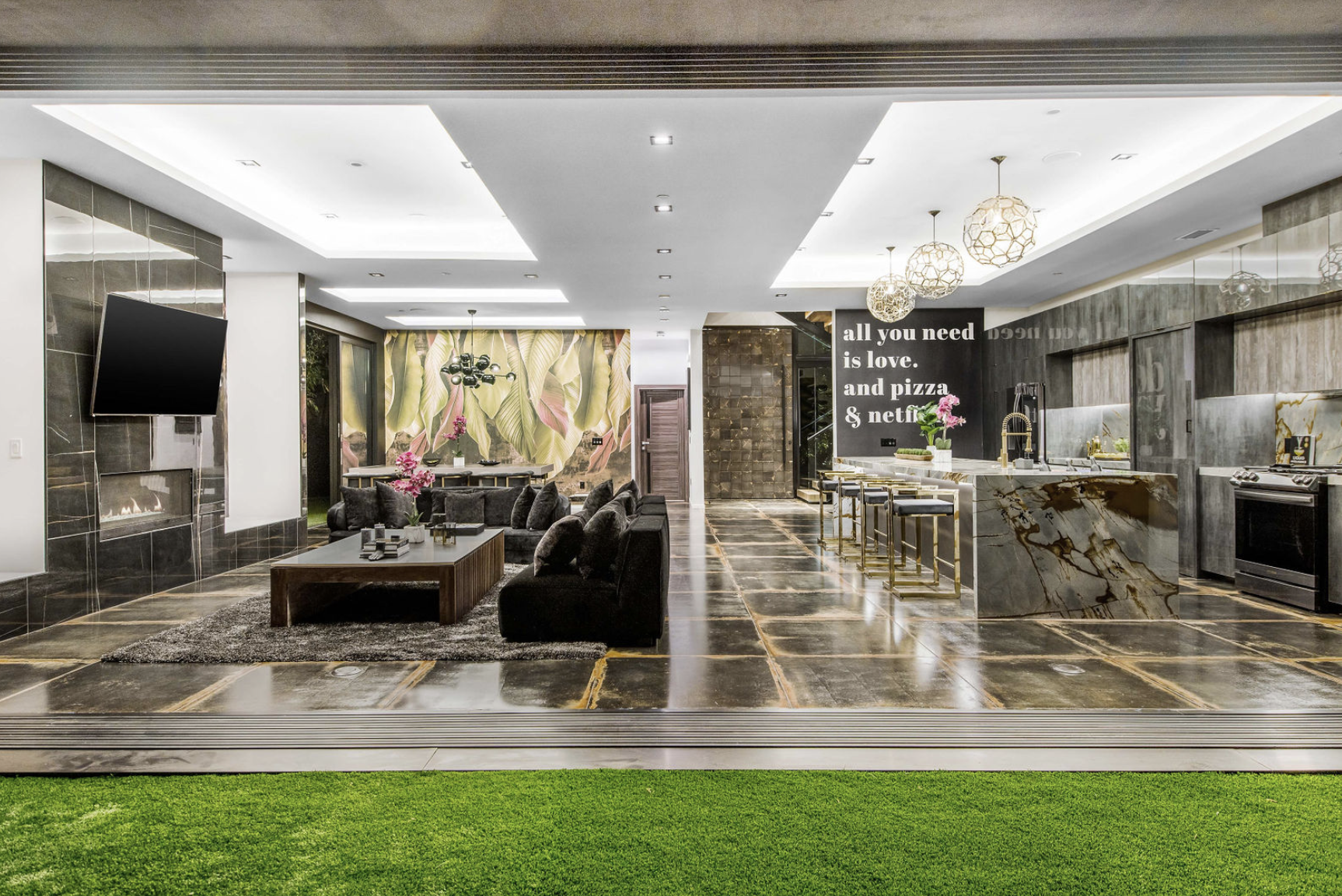 Modern open-concept living room and kitchen with black marble floor, black sofas, colorful wall art of tropical leaves, and white kitchen island with pink flowers and gold accents, illuminated by decorative pendant lights.