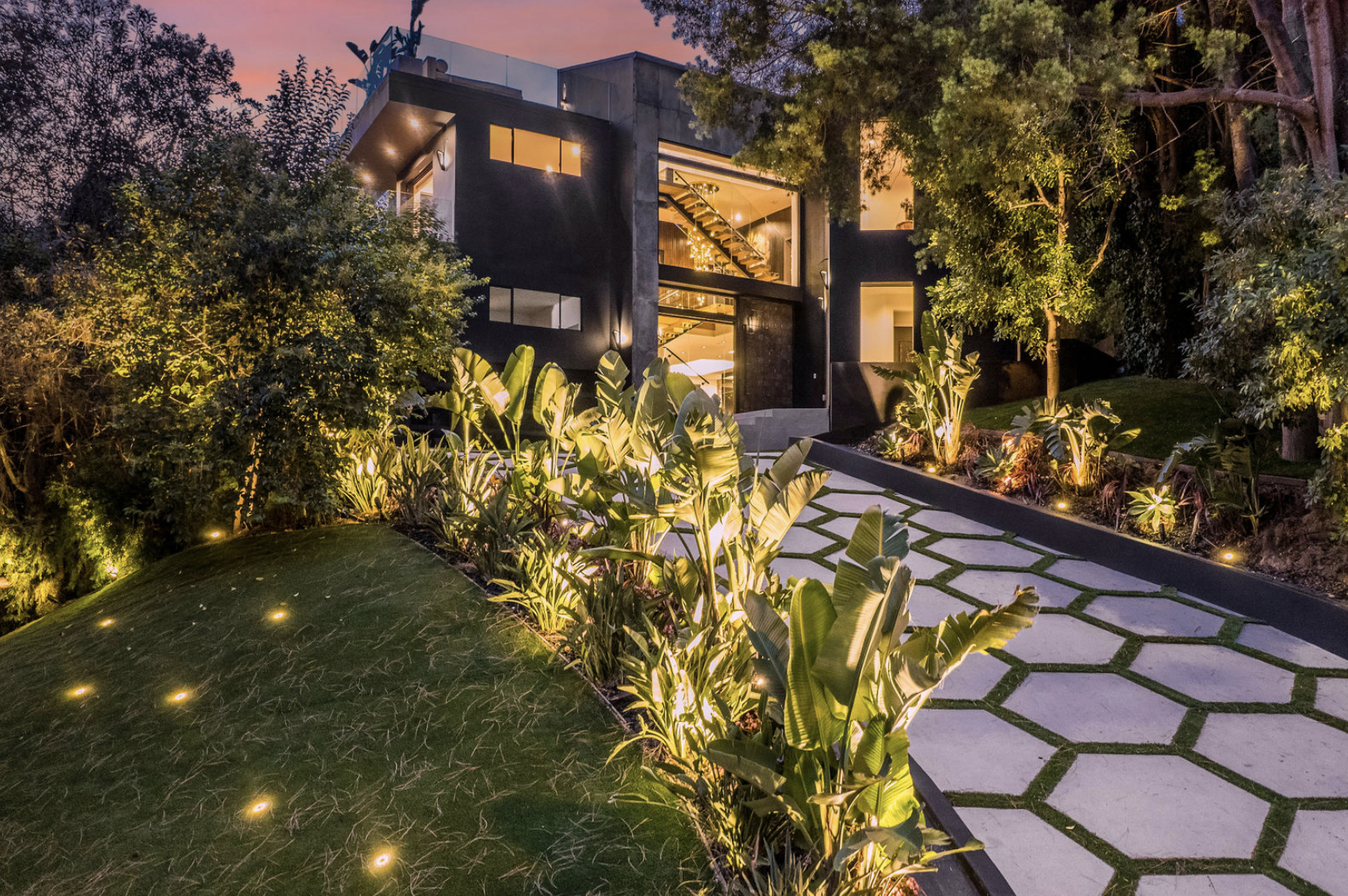 Modern house at dusk with large windows, illuminated staircase inside, surrounded by landscaped garden with pathway and plants, outdoor lighting.