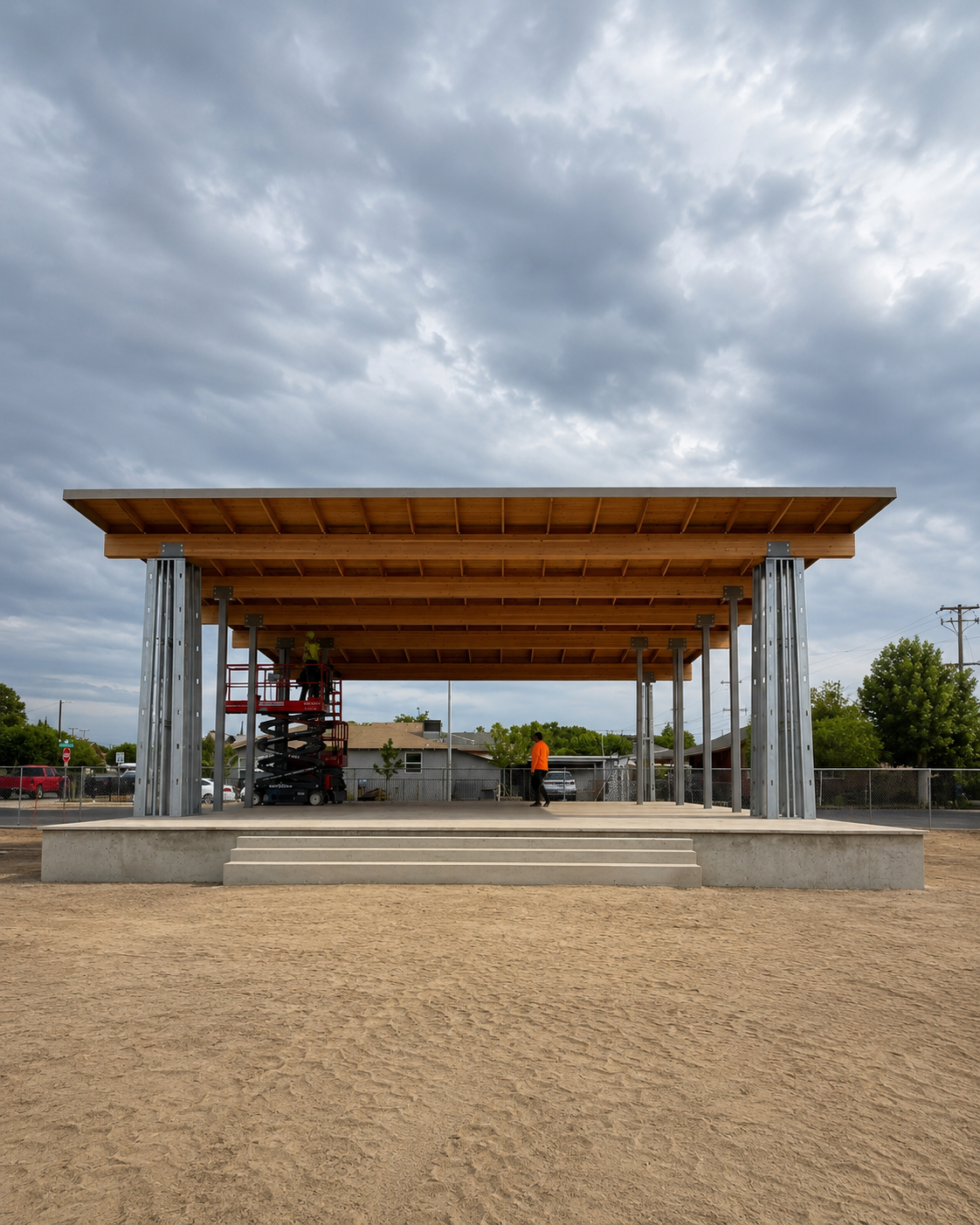 Construction of a large open wooden pavilion with metal supports, situated on a concrete stage, under a cloudy sky, with workers present and surrounding residential area.
