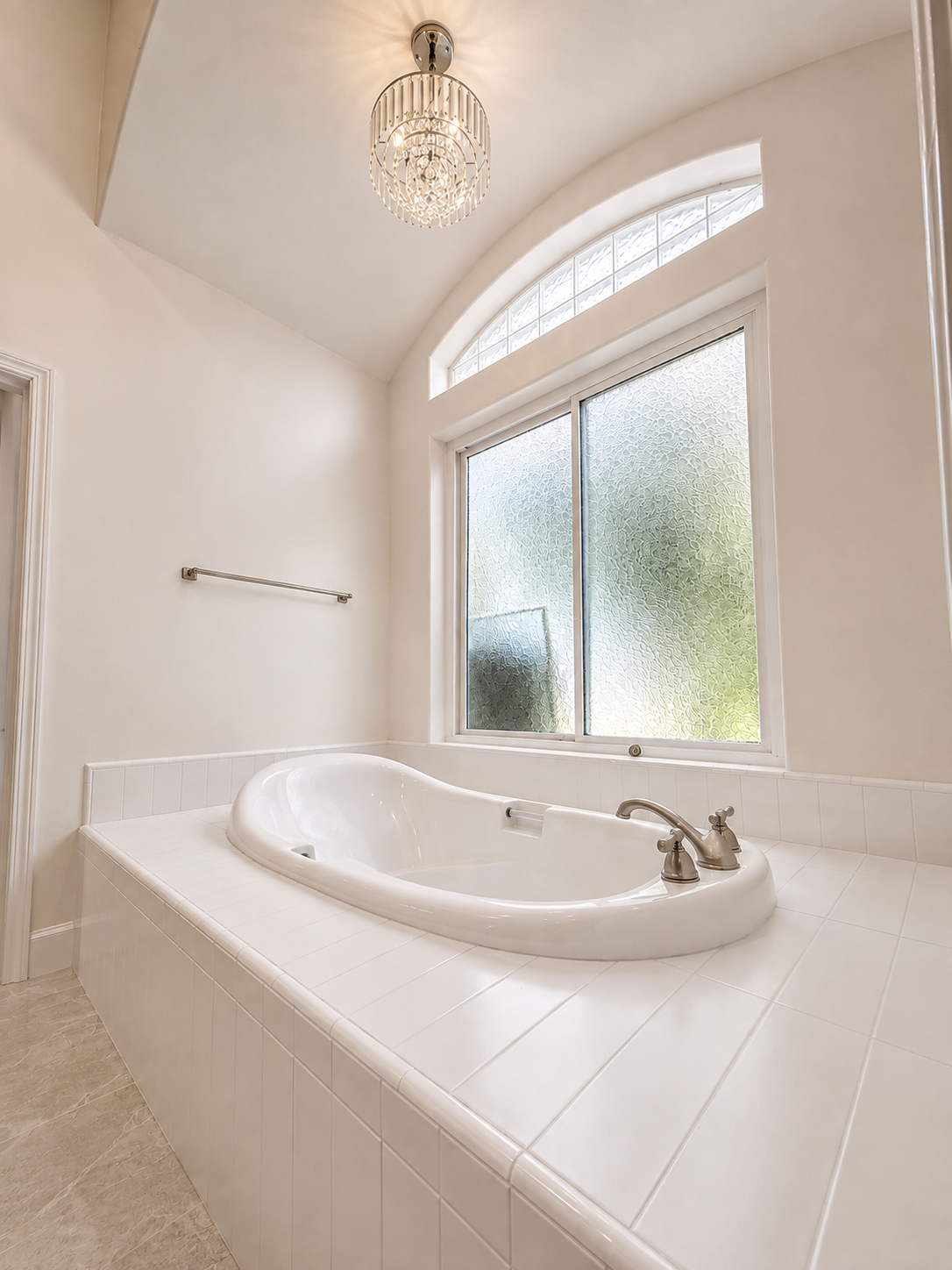 A clean, white bathroom features a built-in bathtub with brushed nickel fixtures, a large frosted window, and a decorative ceiling light fixture.