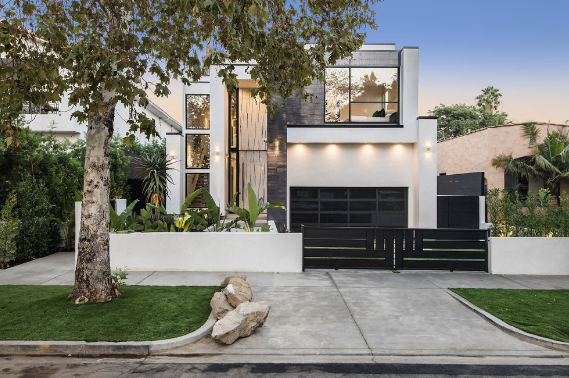Modern two-story house with large windows, surrounded by greenery, a tree in front, and a sidewalk with rocks and grass. The house has a white exterior with black accents, a garage door, and a black gate.
