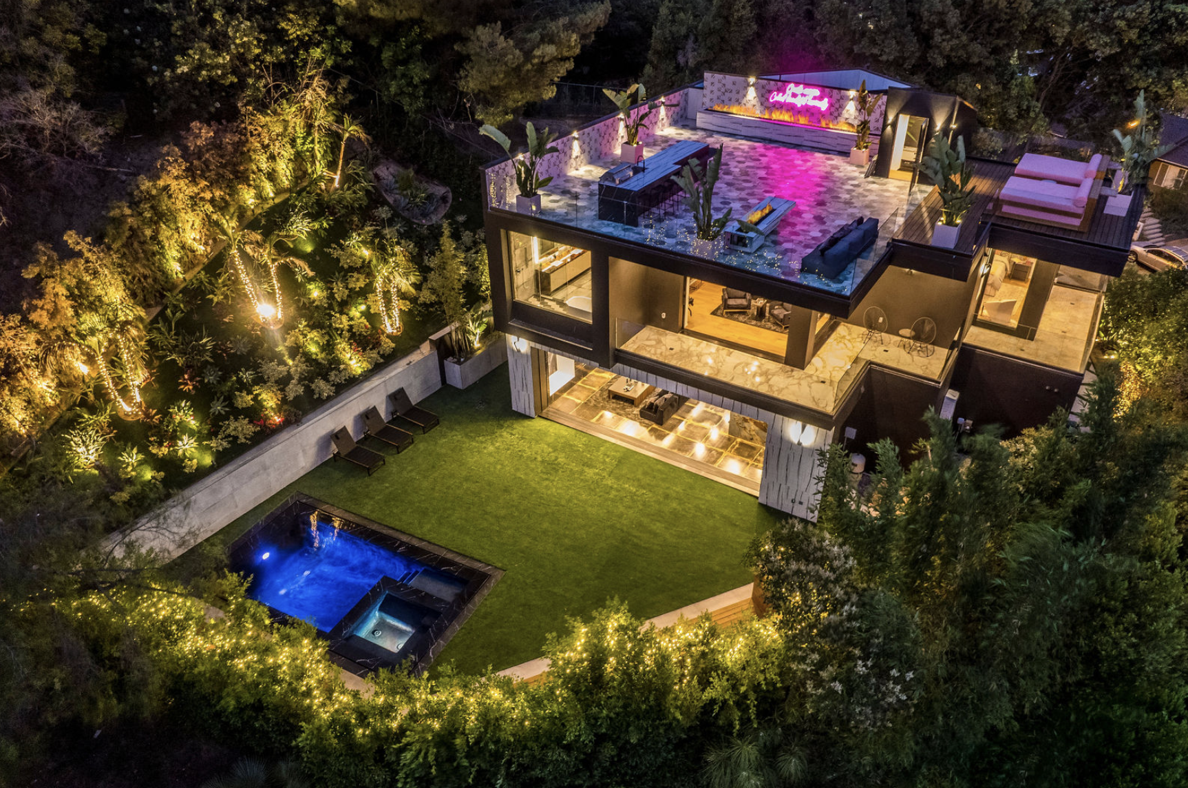 Aerial view of a modern house at night with outdoor lighting, a rooftop lounge area with seating, a pool, a lawn, and surrounding lush trees.