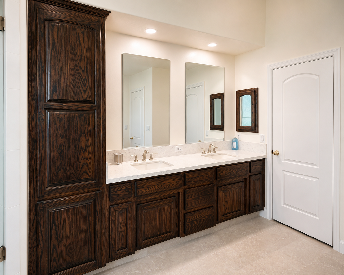 A bathroom double vanity with dark wood cabinets, two sinks with silver faucets, a large mirror, and a small mirror on the side wall. A bottle of hand soap is on the counter. The bathroom has beige tile flooring, recessed ceiling lights, and a white 