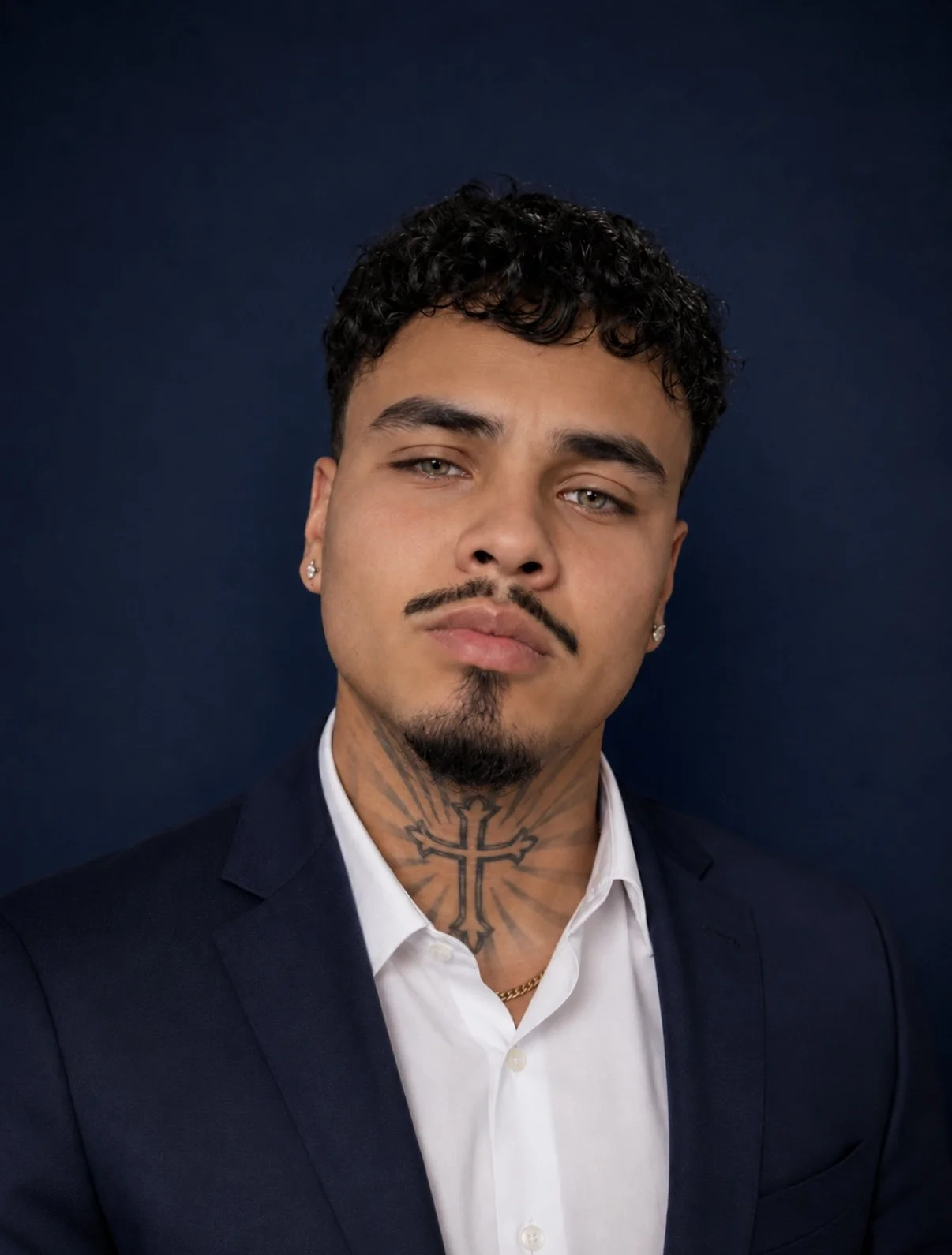 Portrait of a young man with curly black hair, a mustache, and a goatee, wearing a dark suit jacket, white shirt, earrings, and a cross tattoo on his neck, against a dark background.