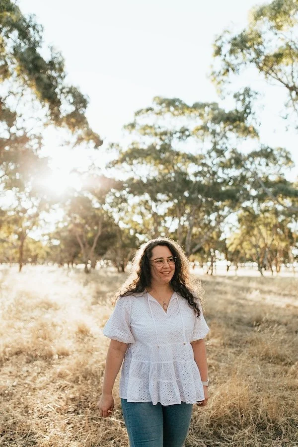 Emily Bryson naturopath standing outdoors in warm golden light wearing a white top and jeans, surrounded by trees