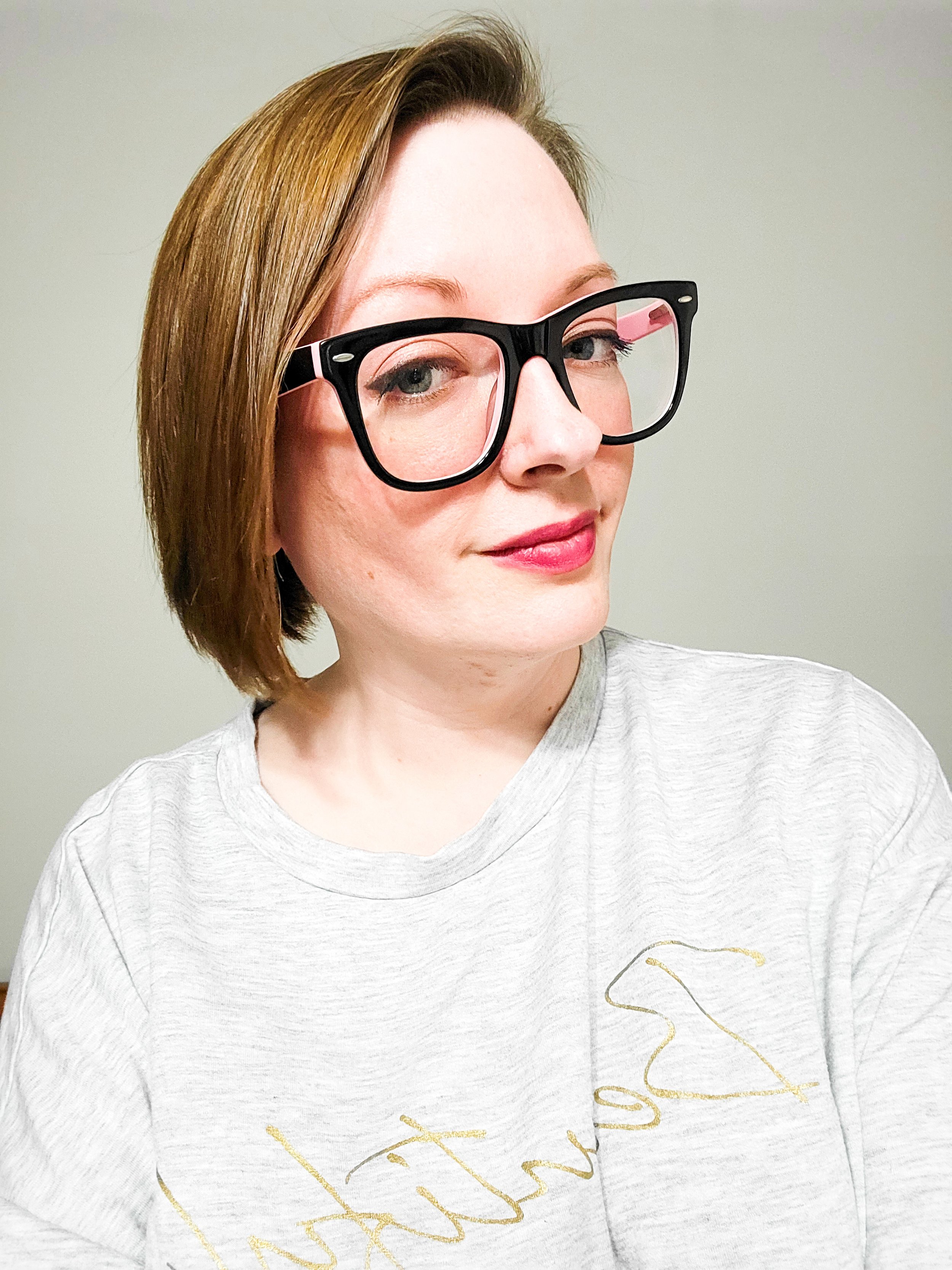 Close-up of a woman with short reddish-brown hair wearing black-rimmed glasses and a light gray T-shirt with gold script writing, standing against a plain light-colored background.