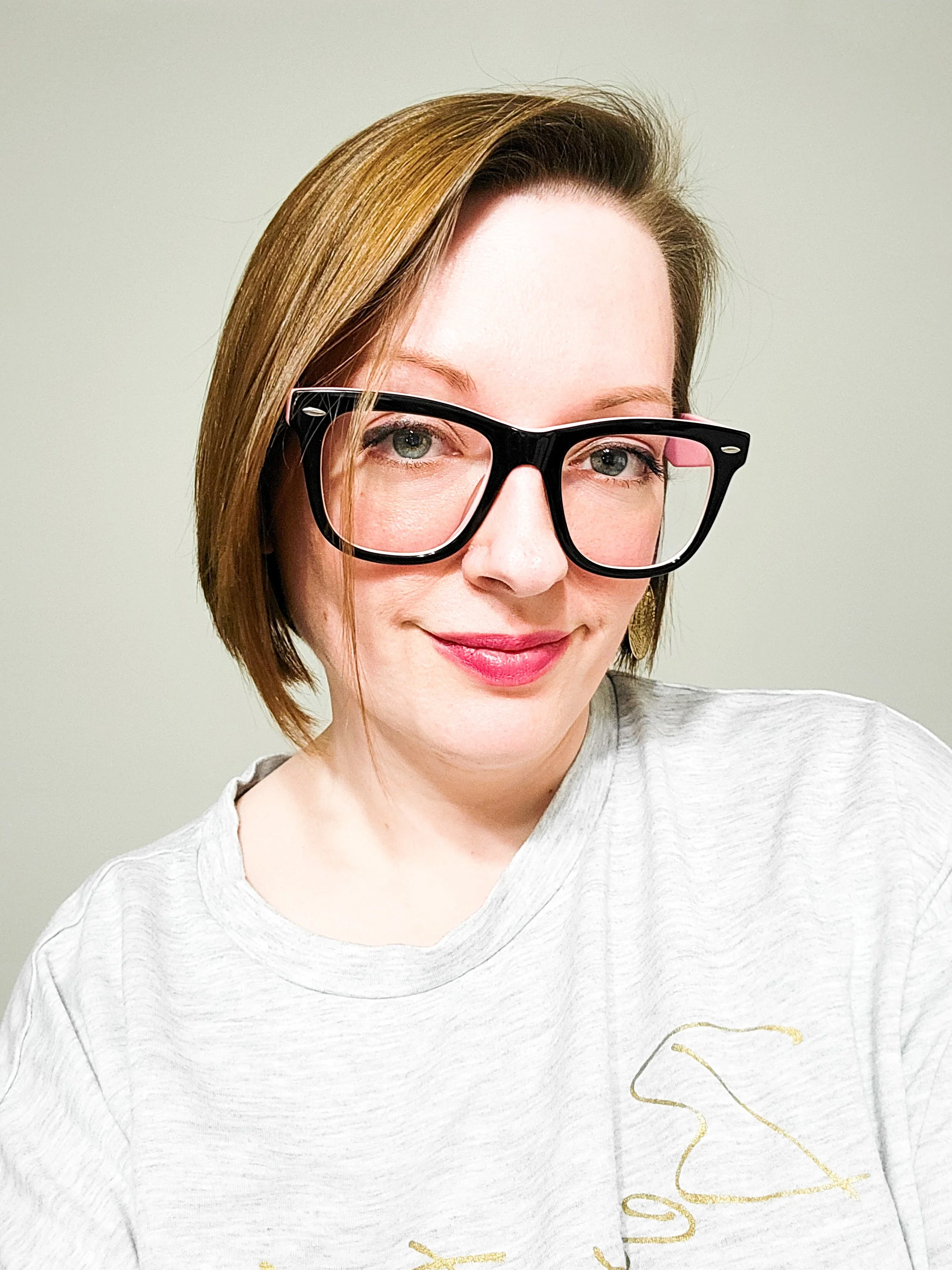 A woman with short light brown hair and glasses, smiling, wearing a gray shirt with a gold design, standing against a plain light-colored background.