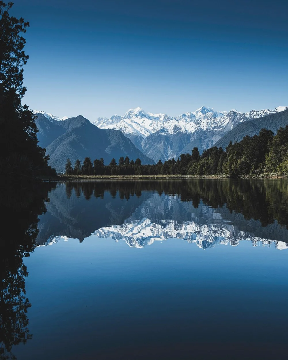 Snow-capped mountains reflected in a calm lake surrounded by green trees under a clear blue sky.