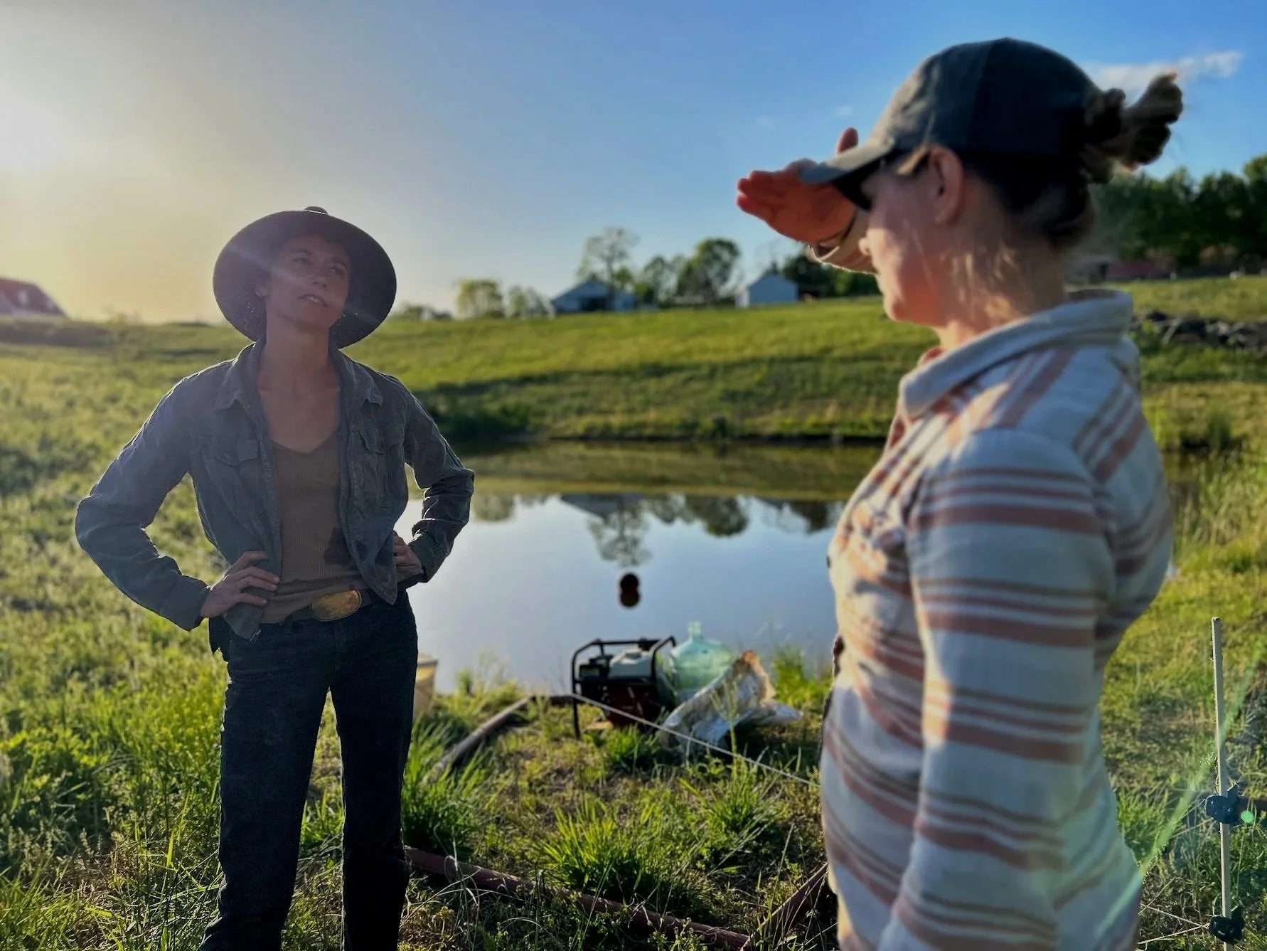 Two women standing outdoors near a pond, with one woman in the foreground wearing a striped shirt and cap, saluting, and the other woman in the background wearing a cowboy hat and denim jacket, talking. The scene is set in a rural area with grassy fields and houses in the distance under a blue sky.