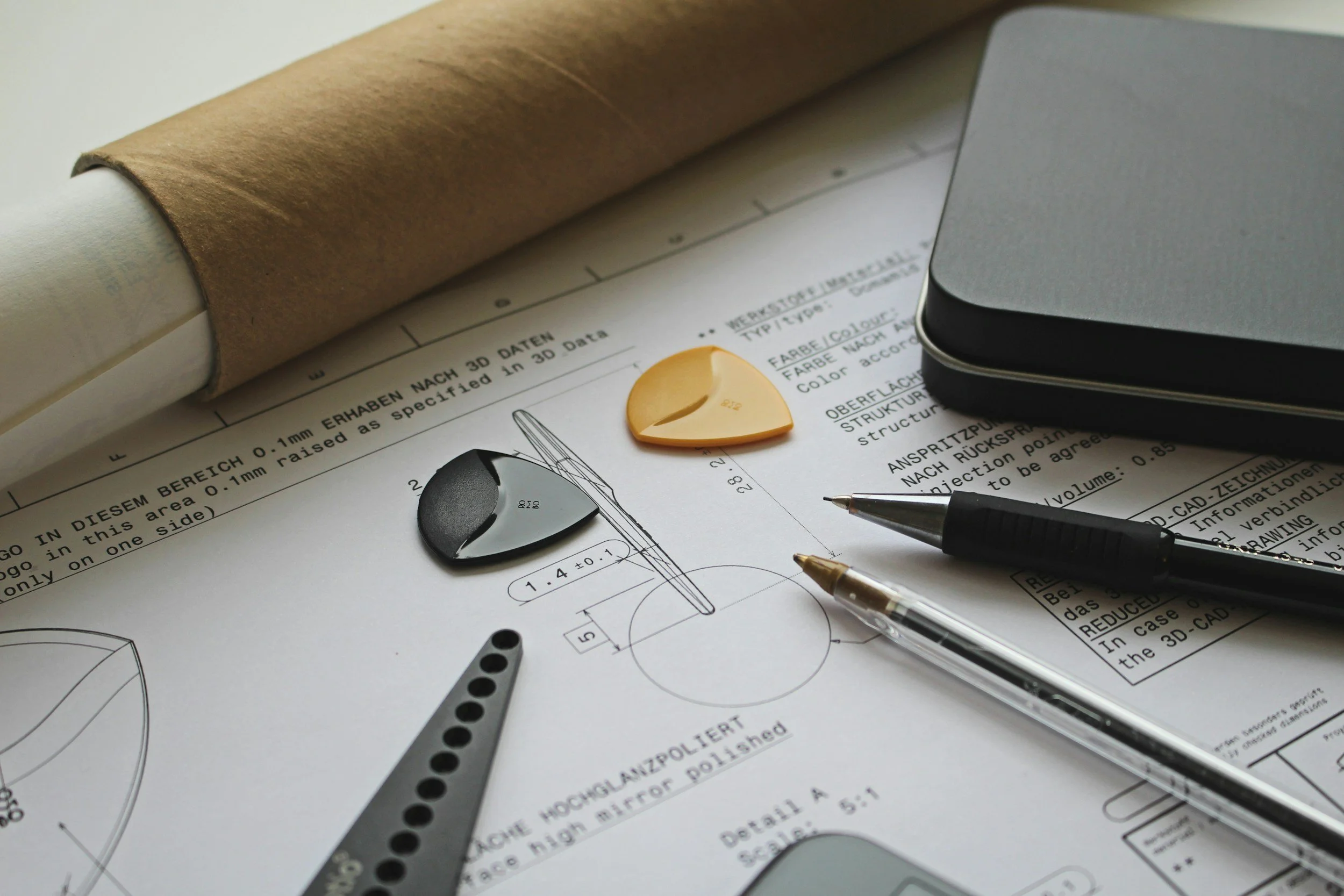 Engineering drawings and tools on paper, including a black pen, a mechanical pencil, a yellow guitar pick, and a metal square ruler, with a roll of brown paper in the background.