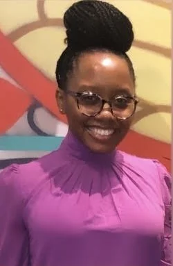 A woman with braided hair, wearing glasses and a purple top, smiling in front of a colorful background.
