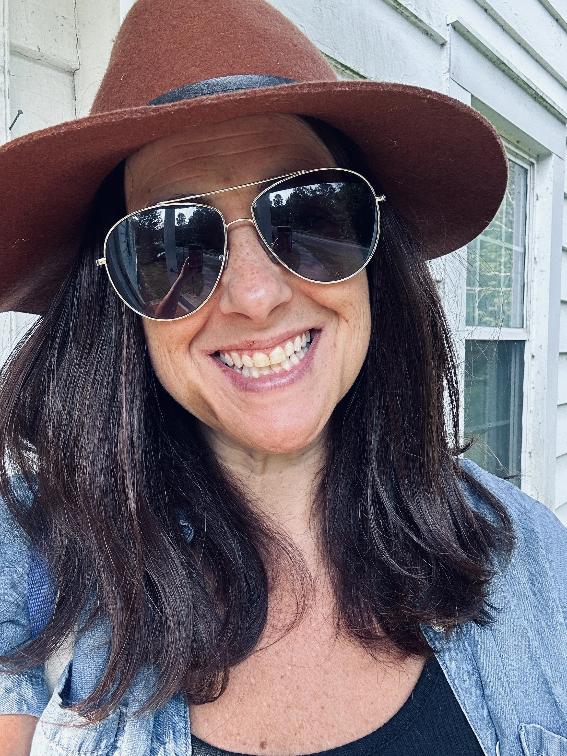 Close-up of a woman smiling outdoors, wearing sunglasses, a wide-brimmed brown hat, a denim jacket, and a black top, with a white house in the background.