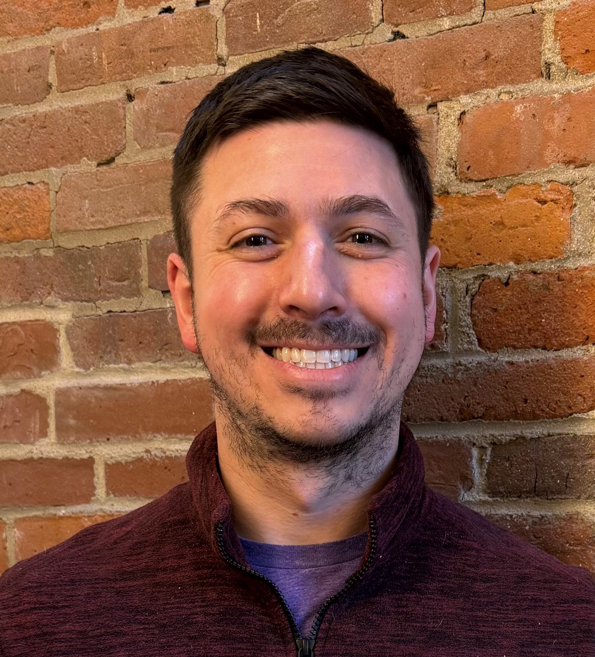 Close-up of a young man's face, showing his forehead, eyes, nose, and smile with a brick wall background.