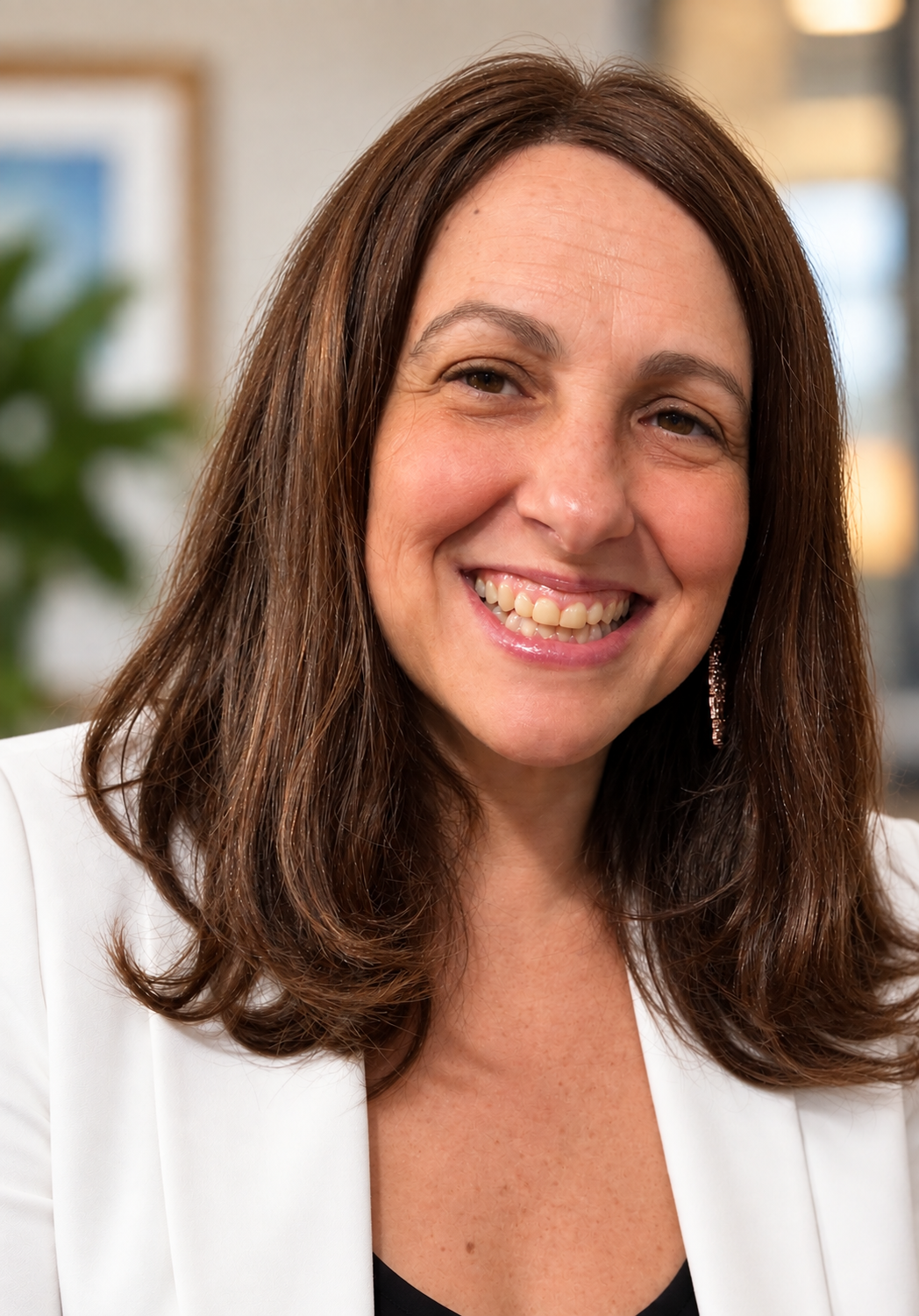 A woman with shoulder-length brown hair, smiling and wearing a white blazer, in an indoor setting.