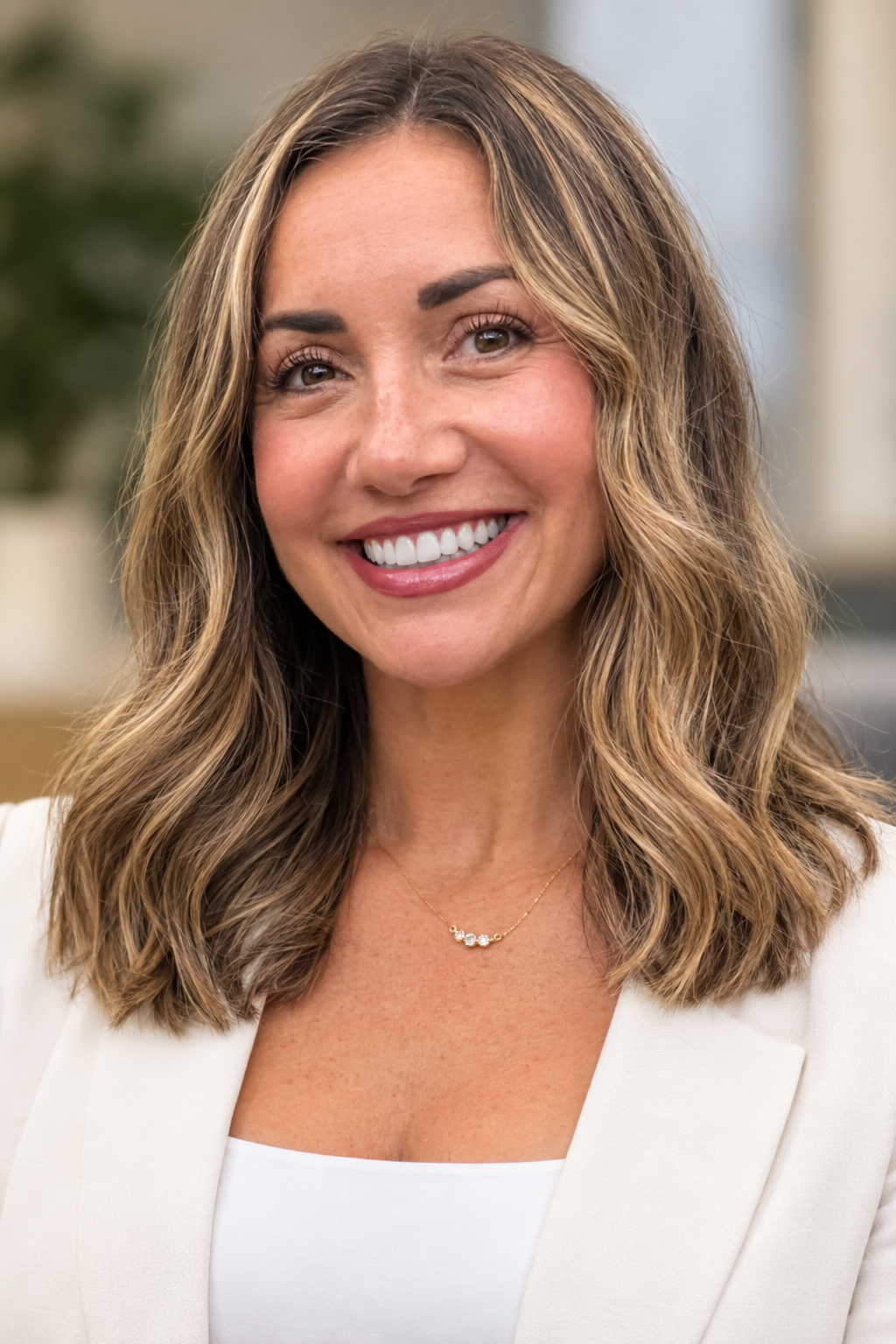 A smiling woman with shoulder-length wavy brown hair with highlights, wearing a white blazer and a delicate gold necklace with small diamonds.