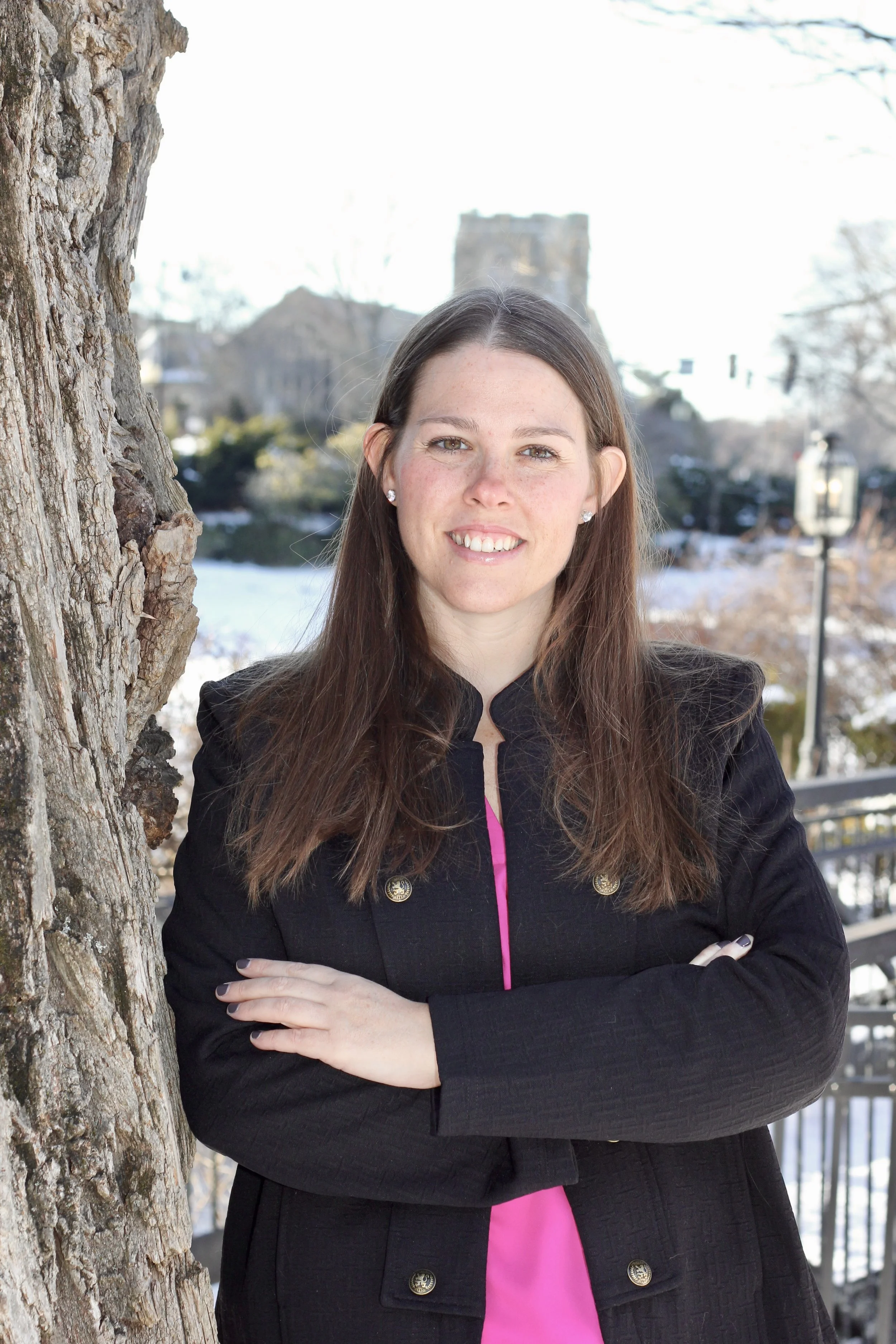 A young woman with long brown hair, wearing earrings and a black jacket over a hot pink top, standing outdoors near a tree, with a castle and snowy landscape in the background.