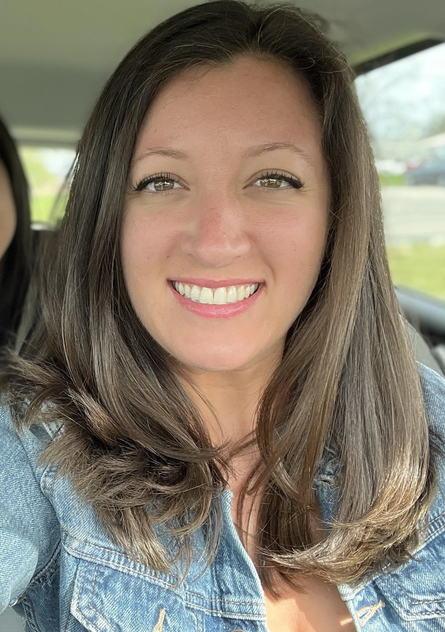 Close-up of a woman with long, wavy brown hair, smiling with white teeth, wearing a light blue denim jacket, sitting in a vehicle with a blurred outdoor background.
