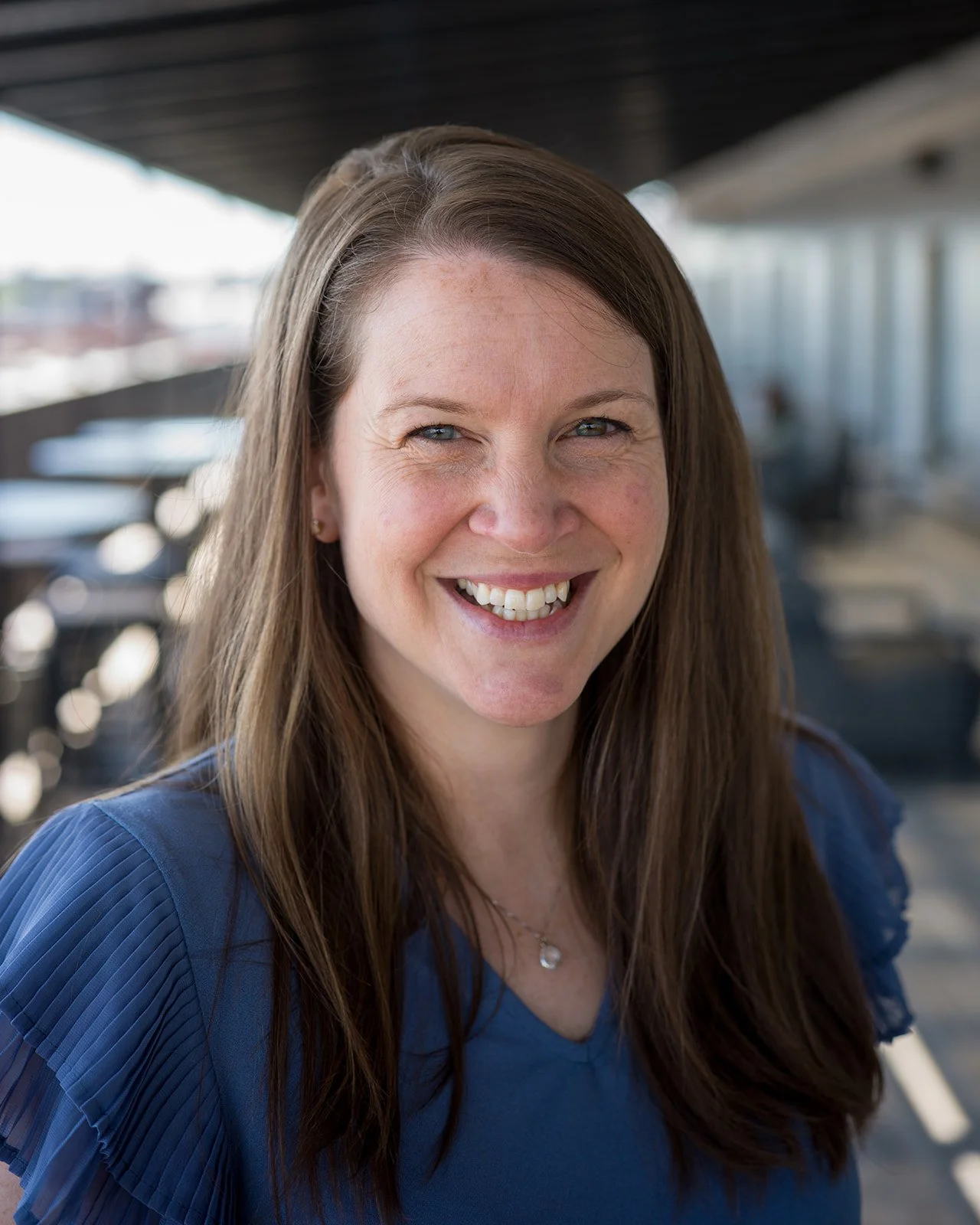 Close-up of a woman with long brown hair smiling at the camera with a background of an outdoor covered patio or balcony.