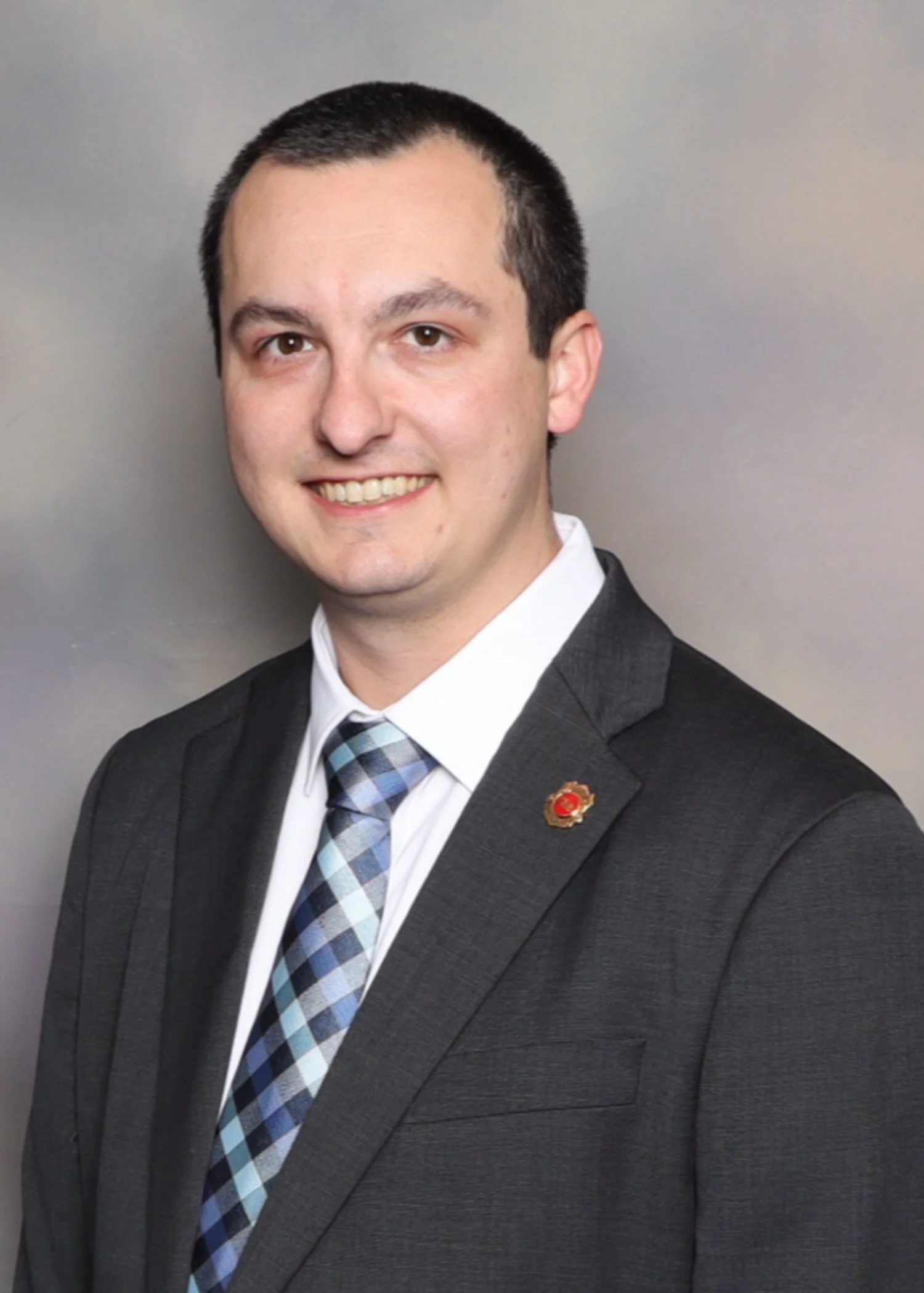 A man in a dark suit, white shirt, and a blue plaid tie, smiling, standing against a plain gray background.