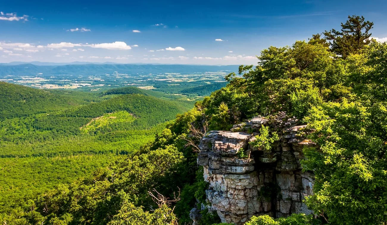 View of a lush green mountainous landscape with a rocky cliff on the right, dense trees, and a distant valley under a bright blue sky with scattered clouds.