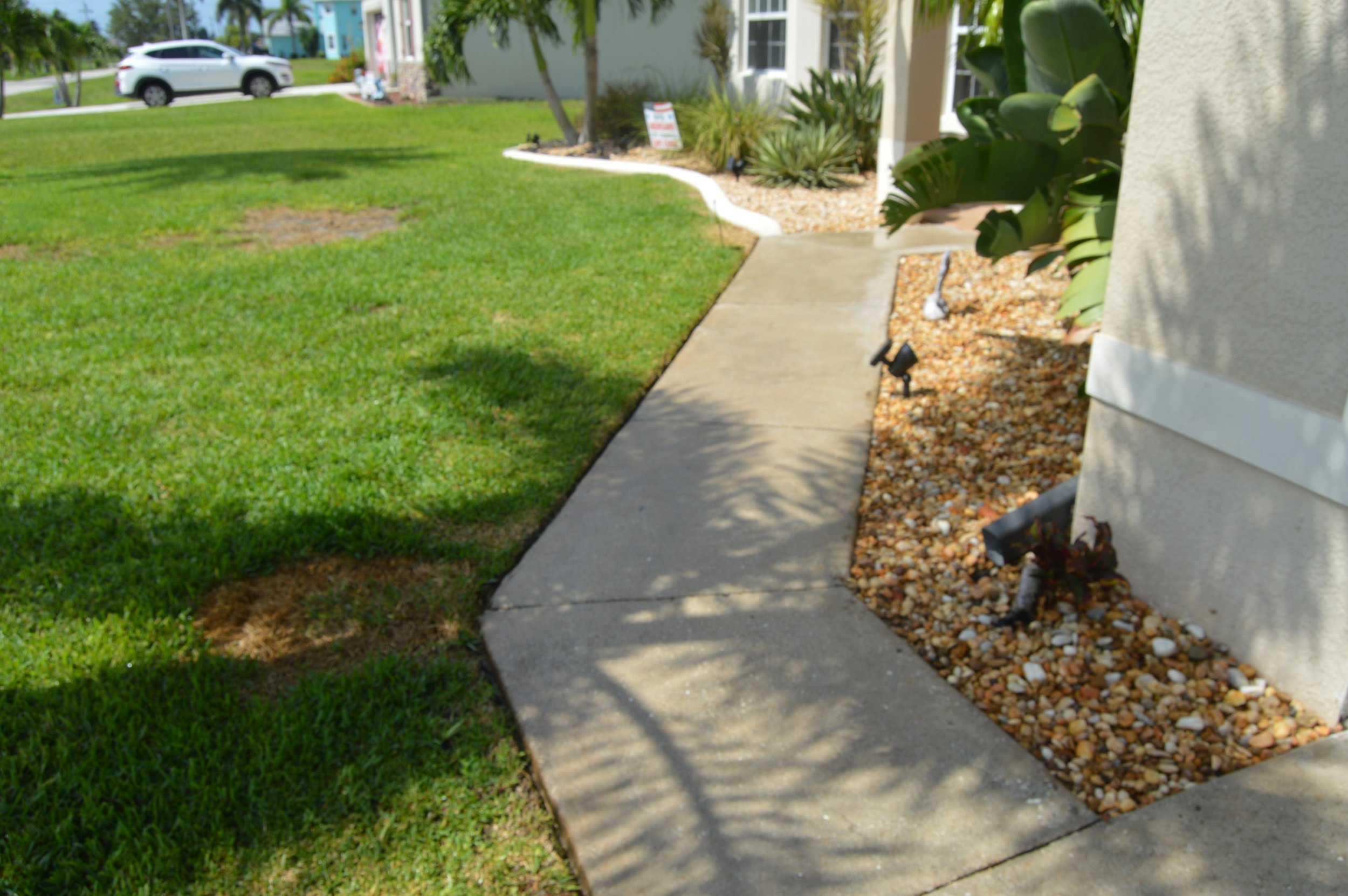 Concrete sidewalk bordered by landscaped garden with small rocks, lush green lawn, and bushes, next to a light-colored building exterior with a security light and pipes.