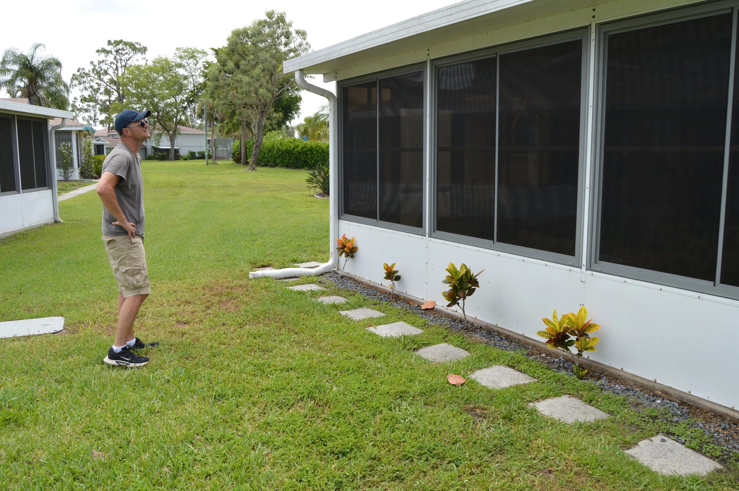 A man wearing a dark baseball cap, sunglasses, a gray t-shirt, and beige shorts stands outside on a grassy lawn assessing soft washing needs for lanai. 