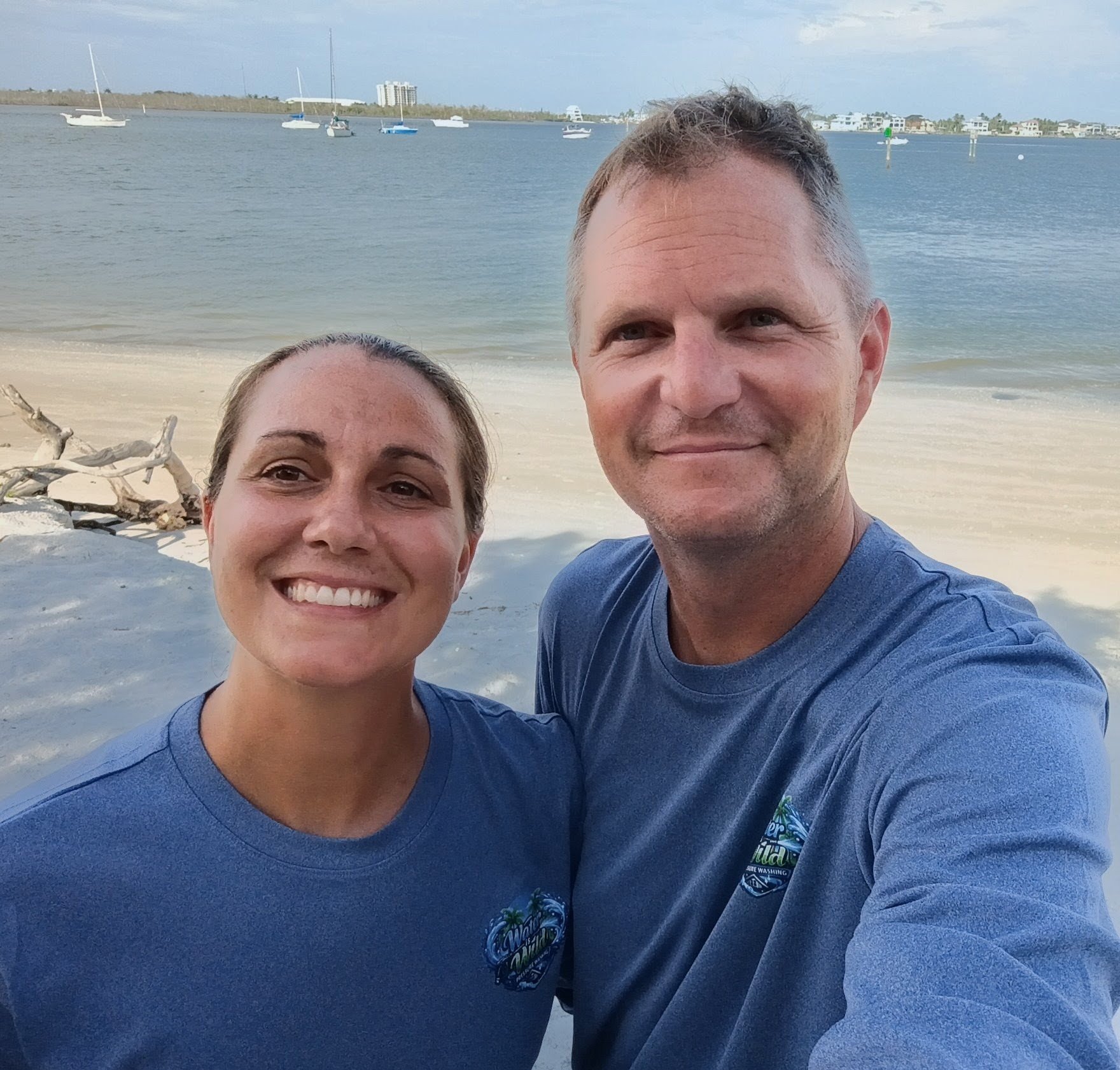 A smiling woman and man taking a selfie at the beach with boats in the water and buildings in the background.