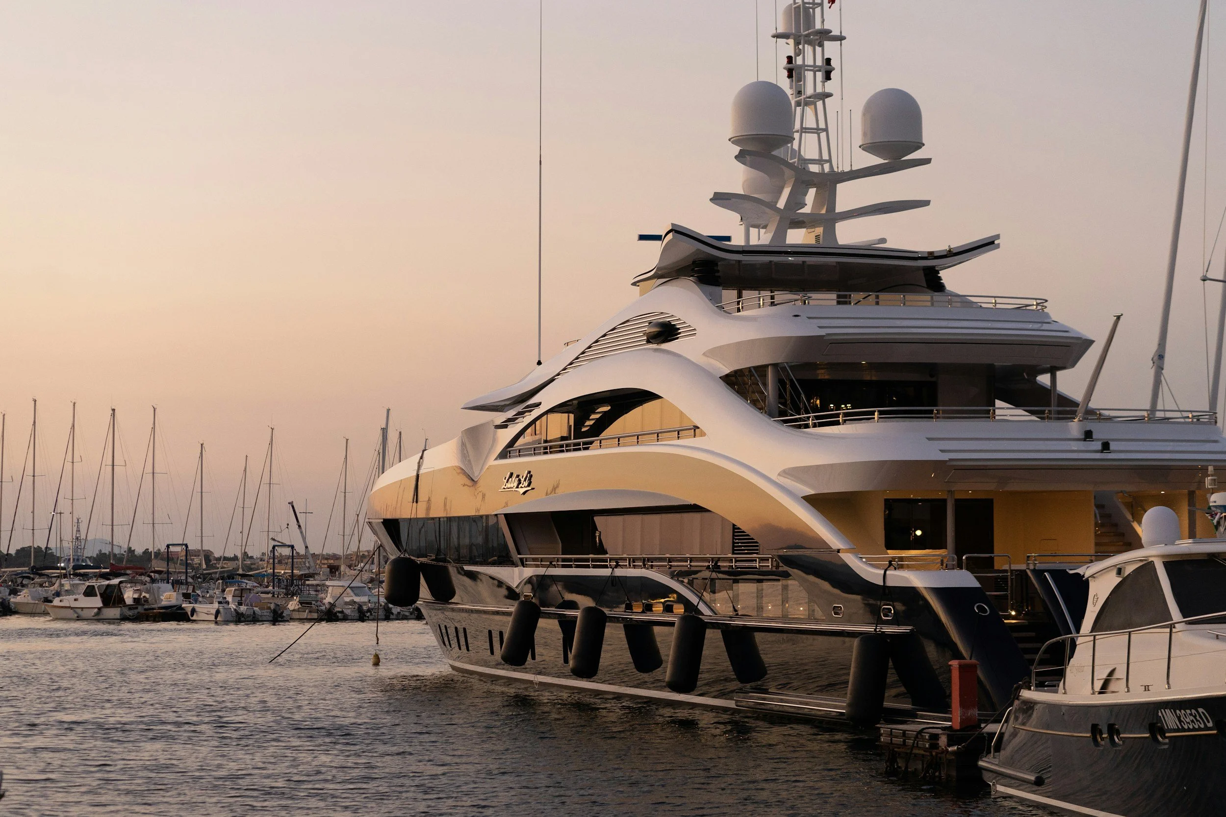 A luxurious yacht docked at a marina with a city skyline and sailboats in the background during sunset.