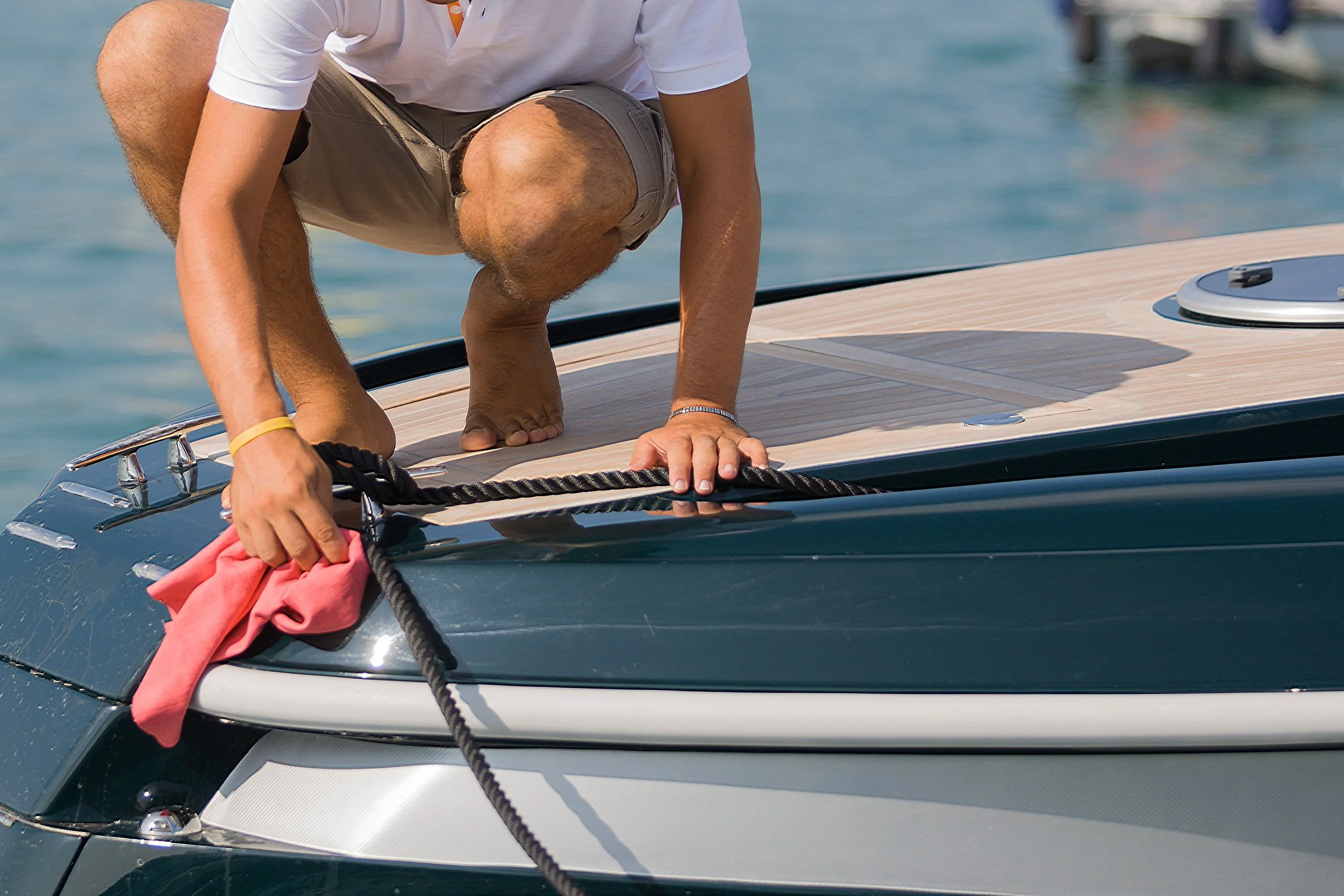 A man is crouched on a boat, reaching out and holding onto a black rope, with a pink cloth attached to it, near the edge of the boat. He is wearing a white shirt, khaki shorts, and a yellow bracelet, with a background of water and a dock.